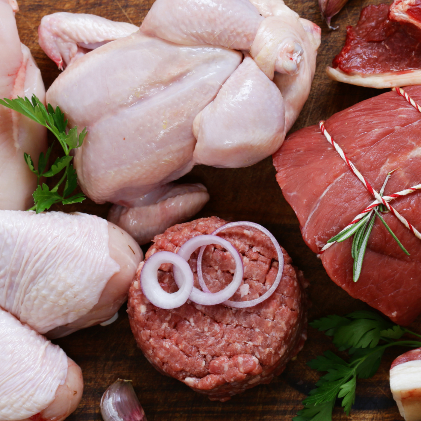 Raw chicken, ground beef with onion rings, and cuts of beef on a wooden surface with herbs and garlic.