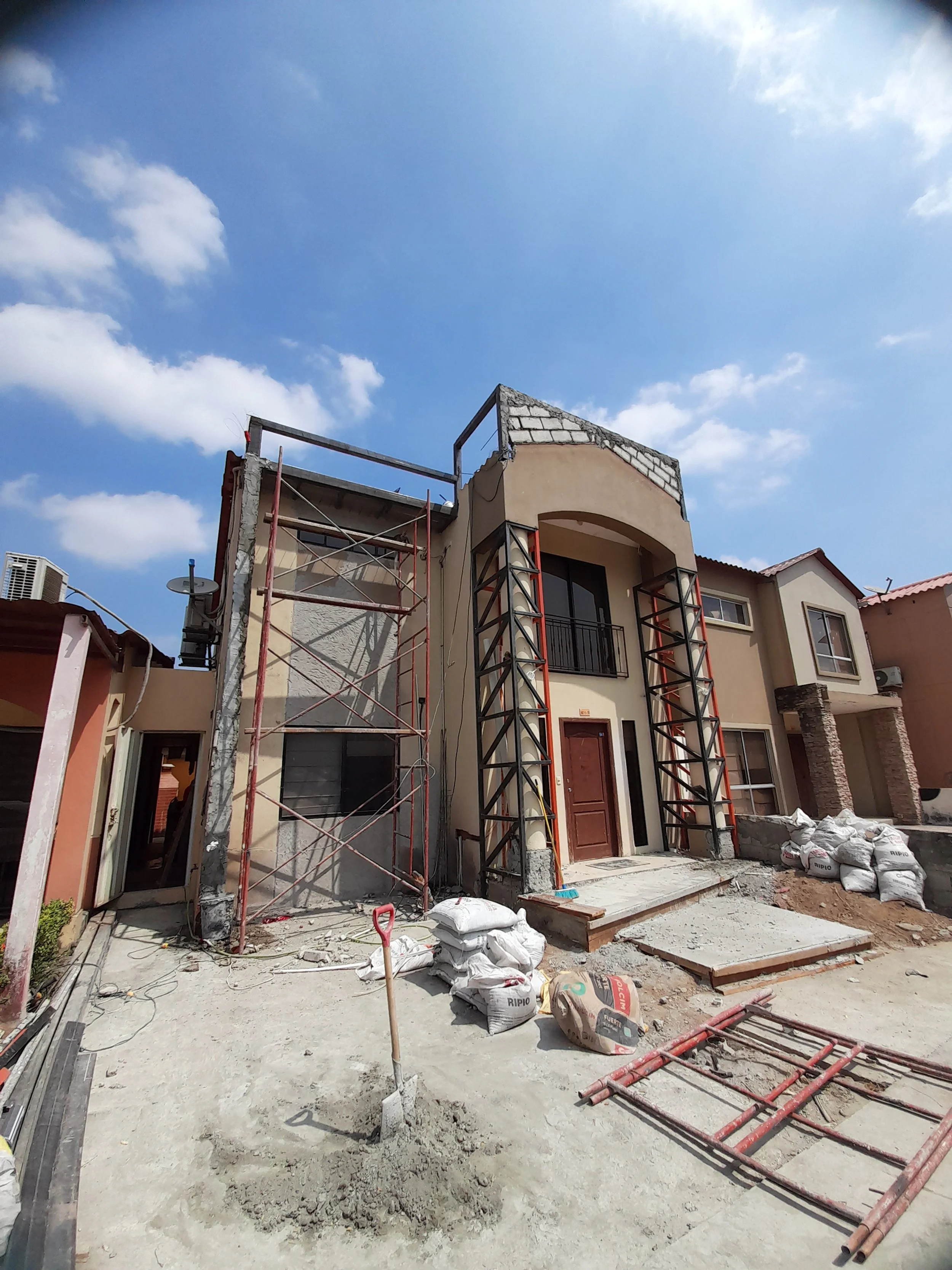 Una casa en construcción con andamios y sacos de cemento en el frente, en un día soleado con cielo azul y algunas nubes.