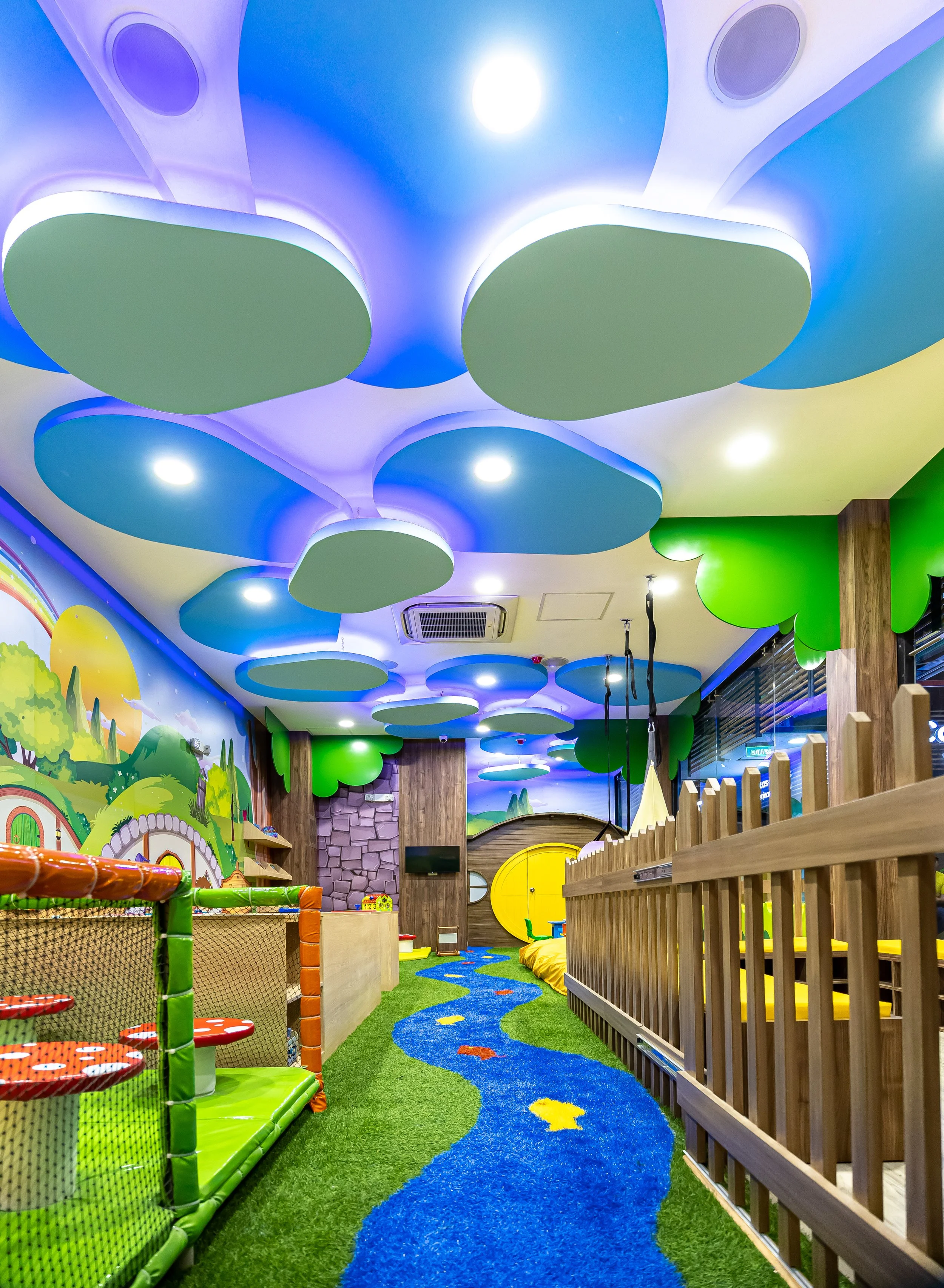 Sala de juegos para niños con decoración de naturaleza y nubes en el techo, pared con paisaje y puerta redonda amarilla.