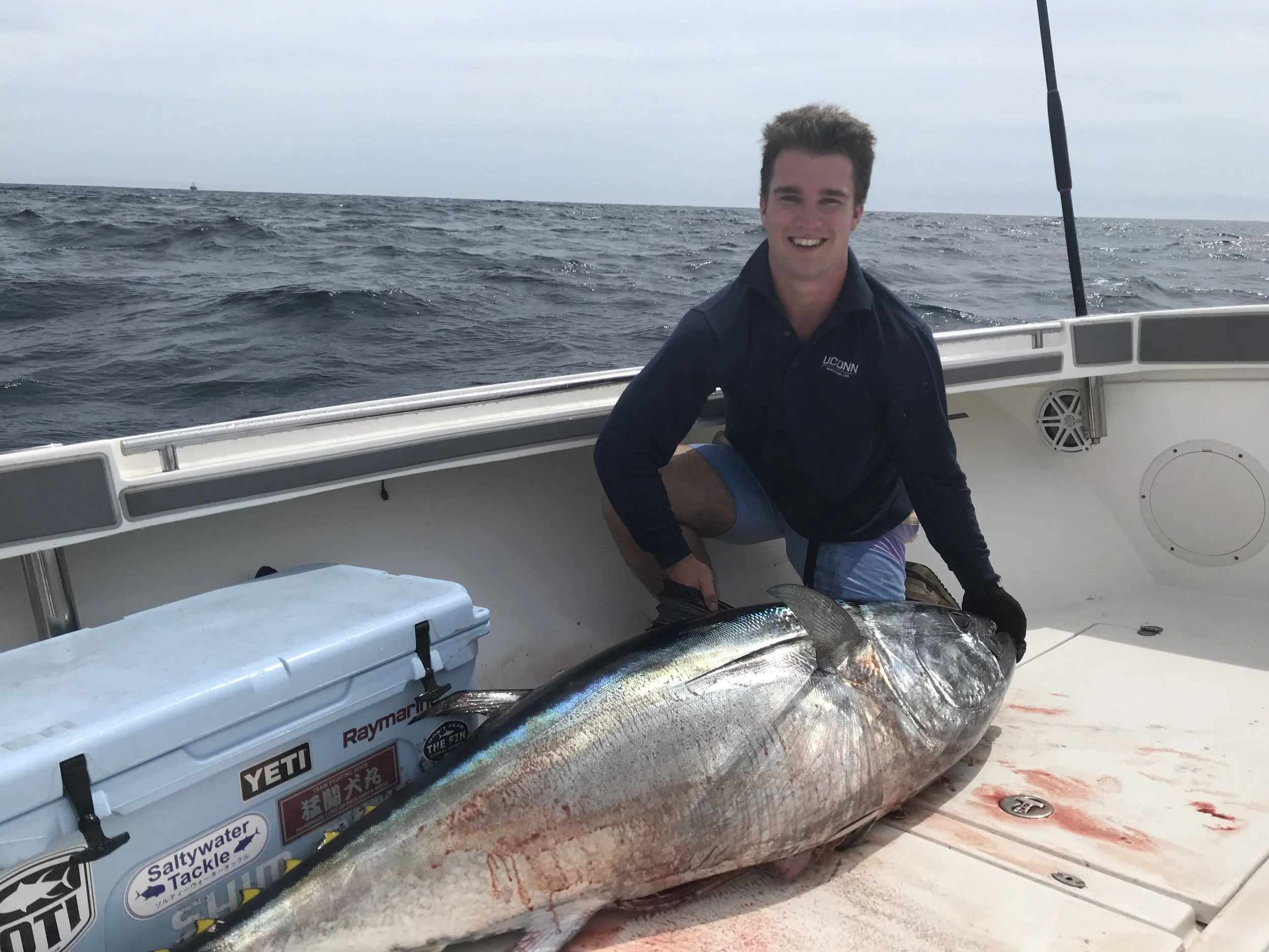 A young man on a boat holding a large fish, with the ocean in the background, smiling at the camera.