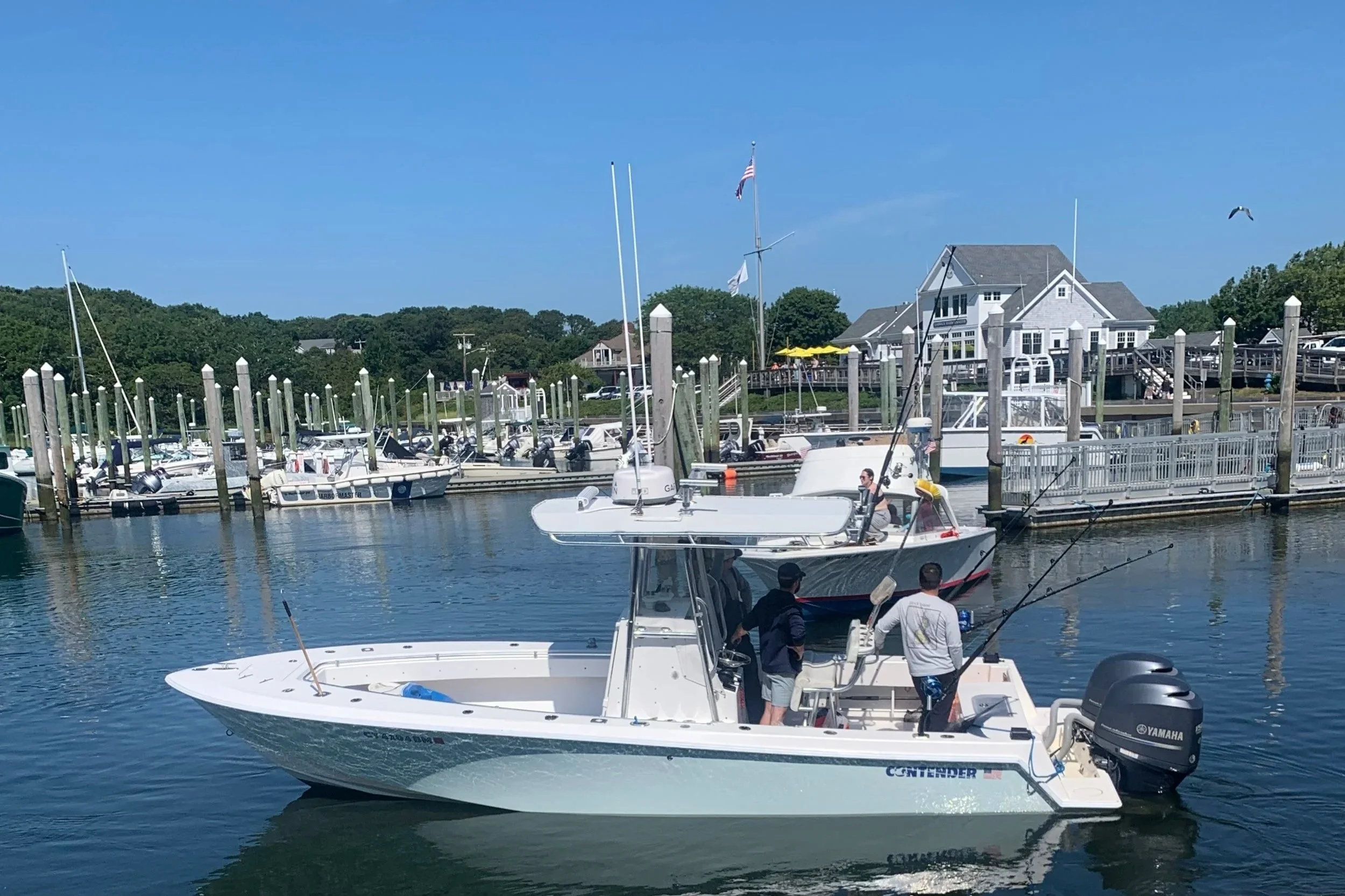 A marina with boats docked, including a white powerboat with an outboard engine and a smaller sailboat with a mast and American flag, under a clear blue sky.