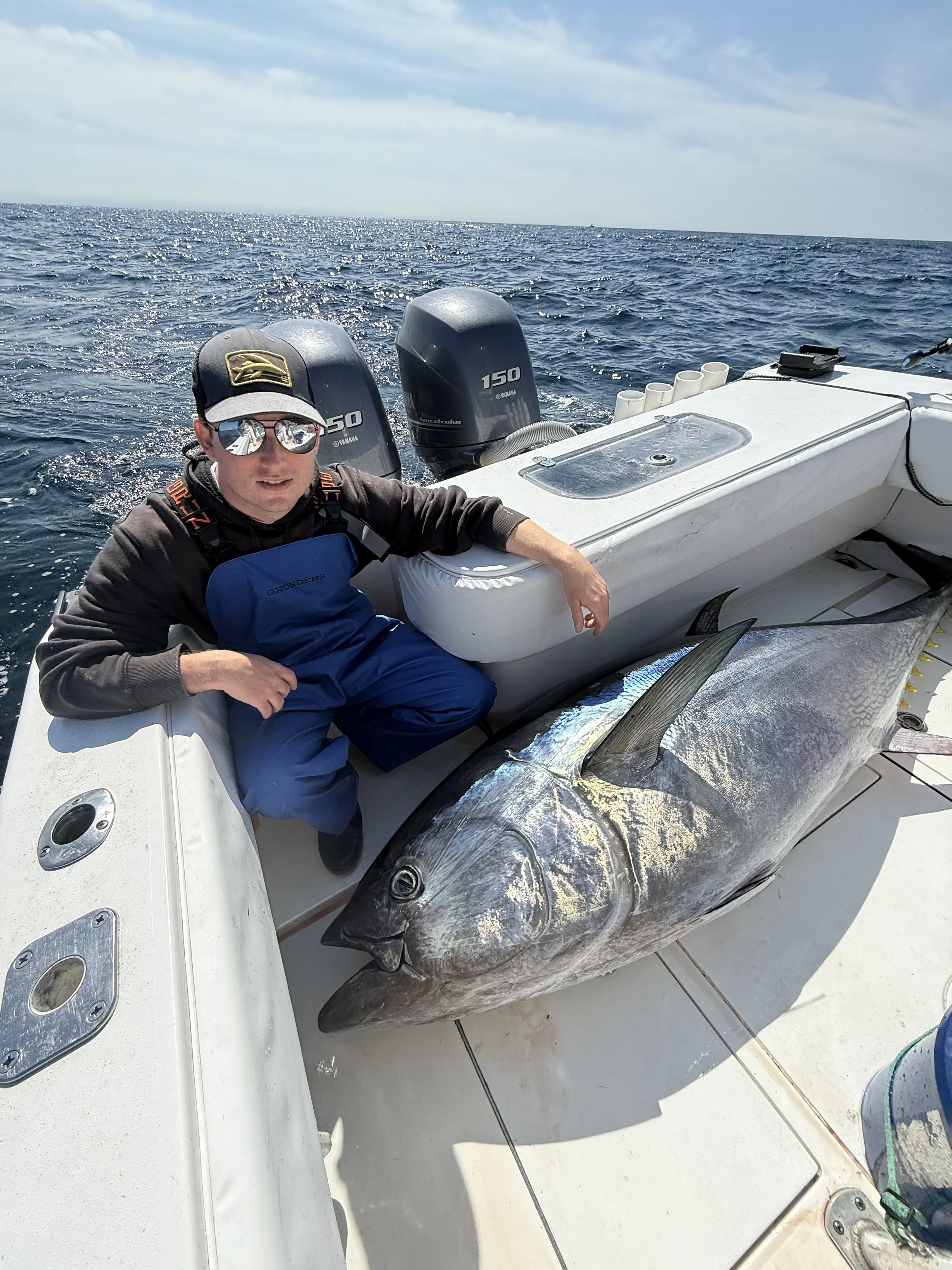 A person on a boat squatting next to a large fish, possibly a tuna, on the deck with the ocean in the background.