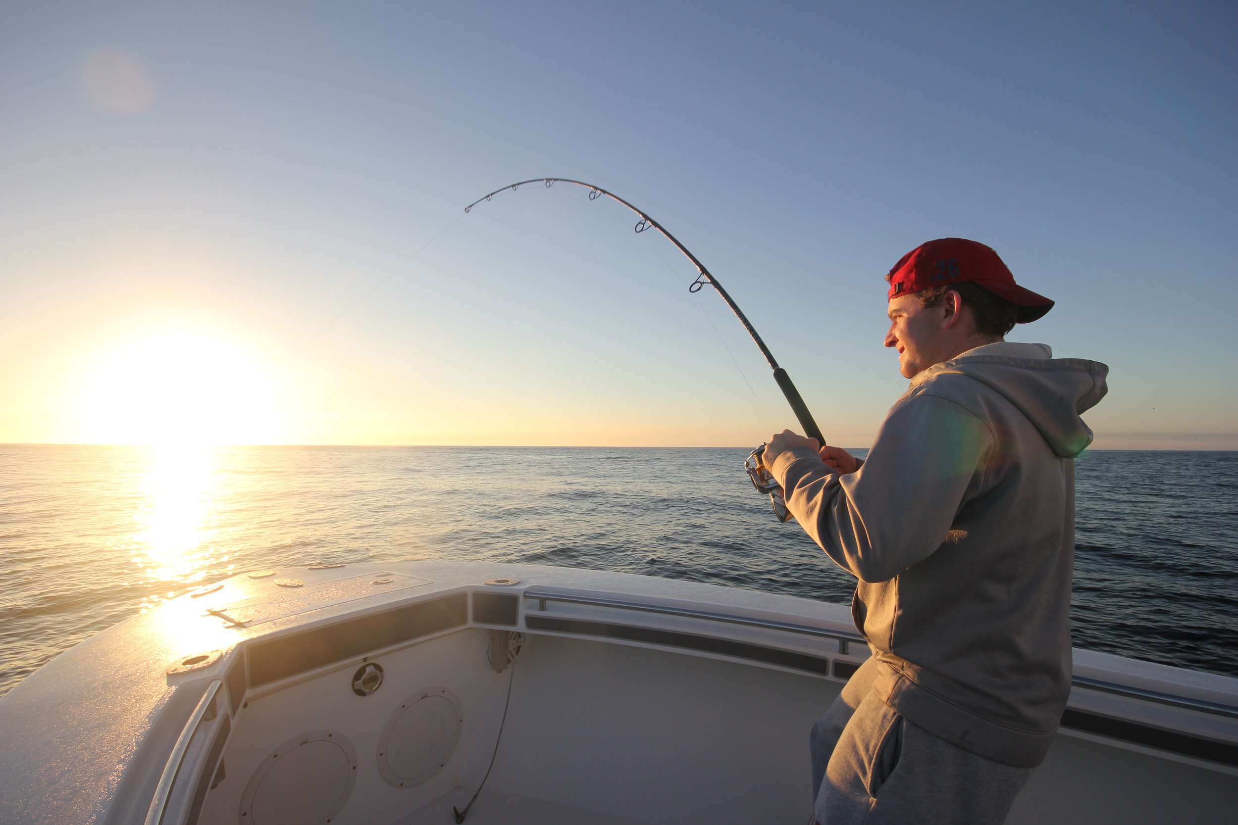 A young man wearing a grey hoodie and a red cap fishing from a boat at sunset.