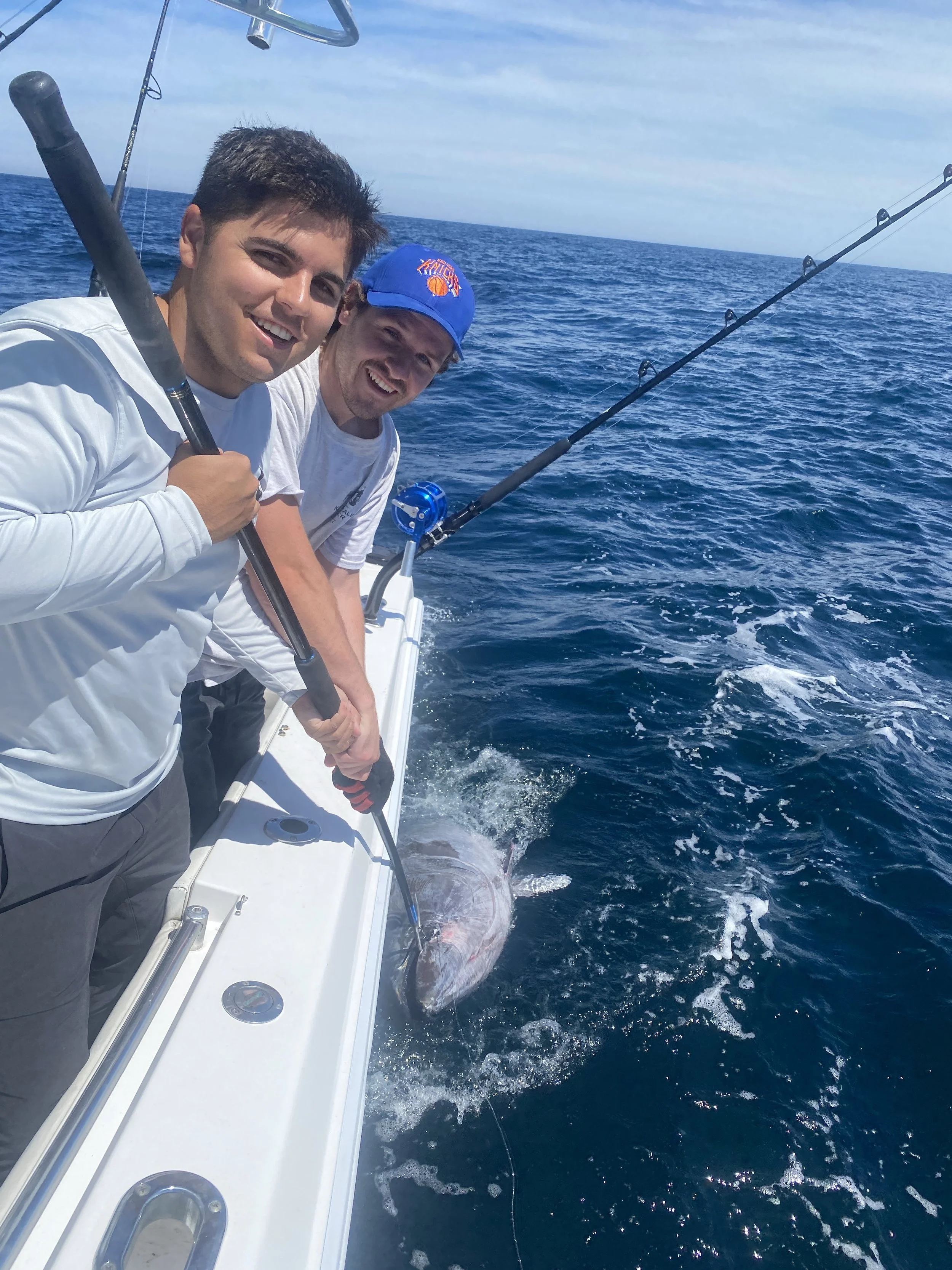 Two men on a boat holding a fishing rod with a large fish caught, on the ocean under a partly cloudy sky.