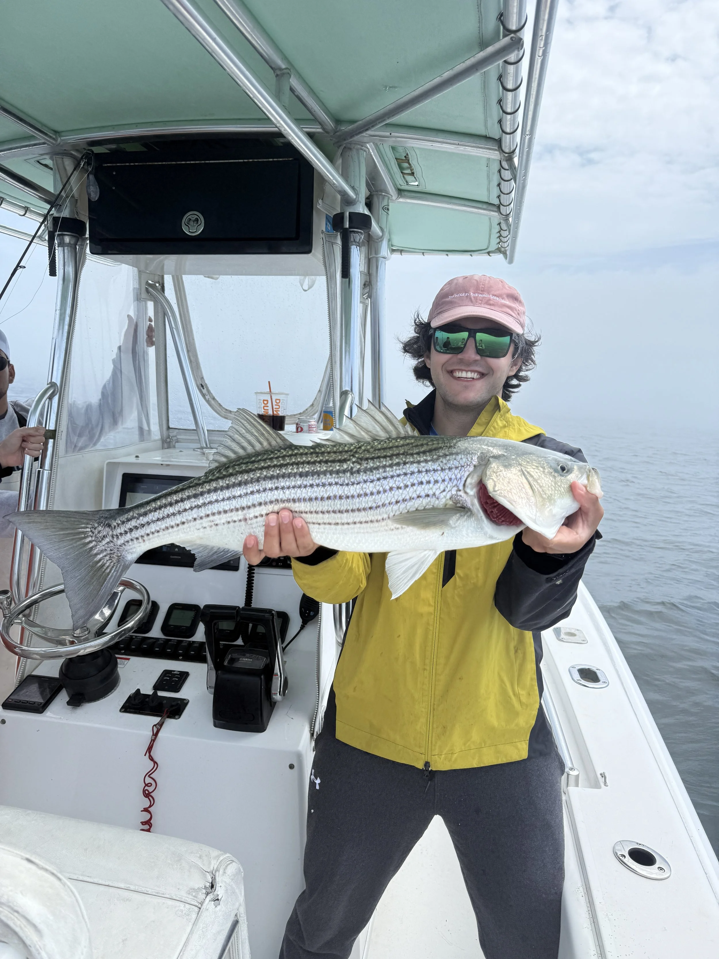 A man in a yellow jacket and sunglasses holding a large striped fish on a boat.