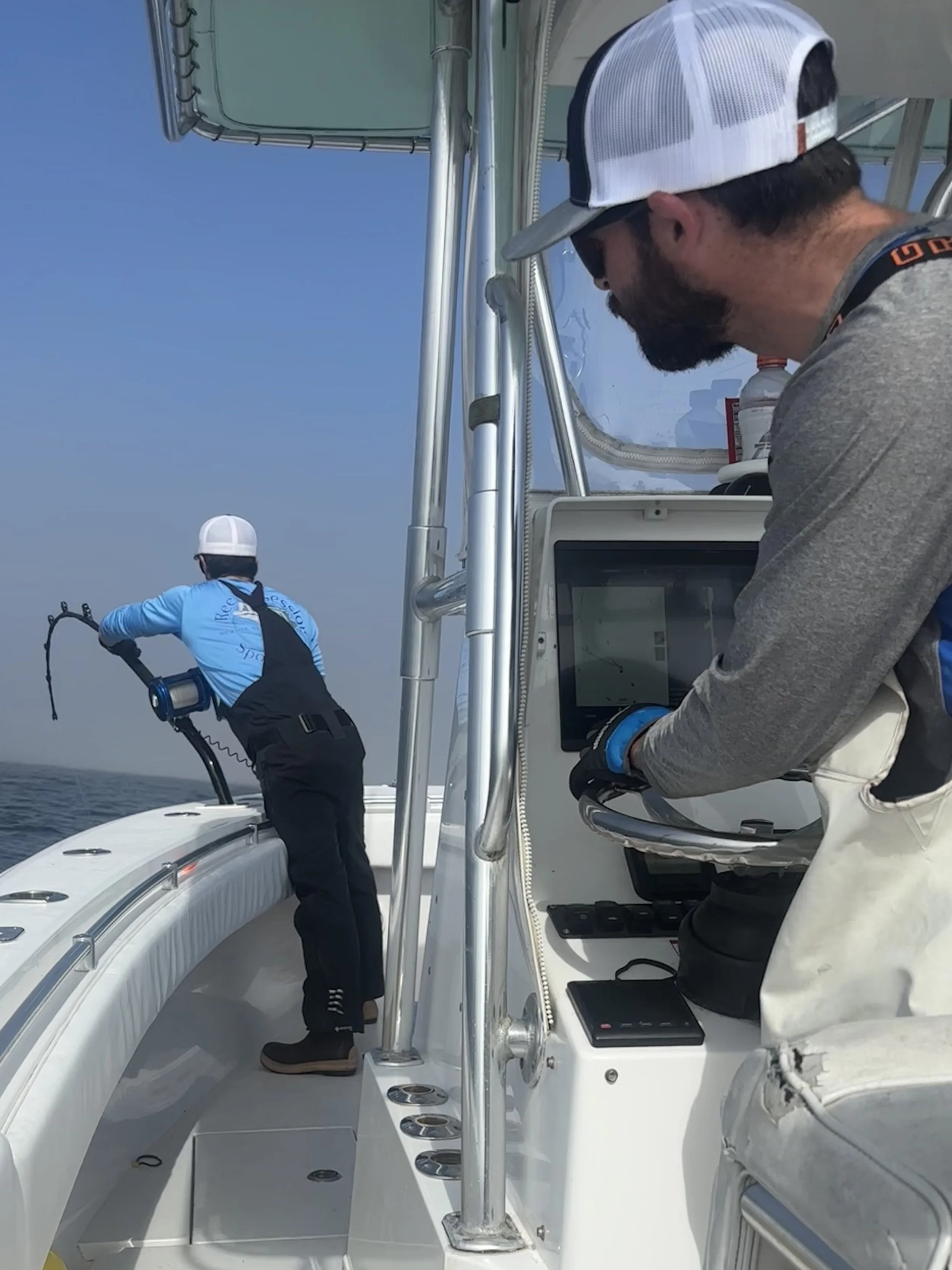 Two men on a boat, one at the wheel looking at a screen, the other in the back using a sonar device to scan the water.