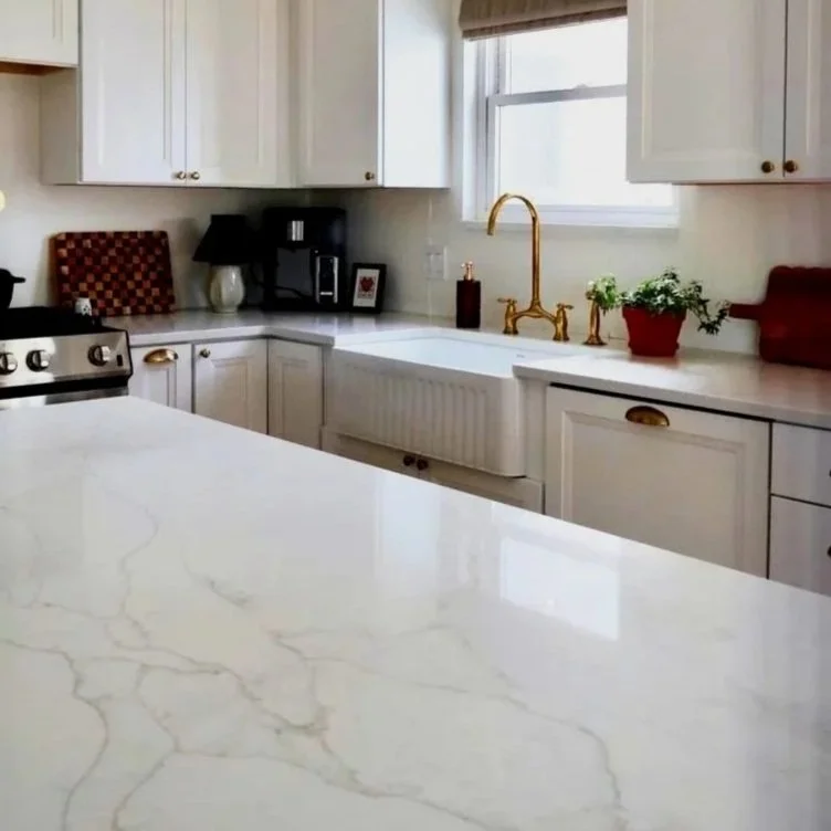 A bright kitchen with white cabinets and a marble island, gold faucet, window above the sink, coffee maker, decorative plants, and some small appliances on the counter.