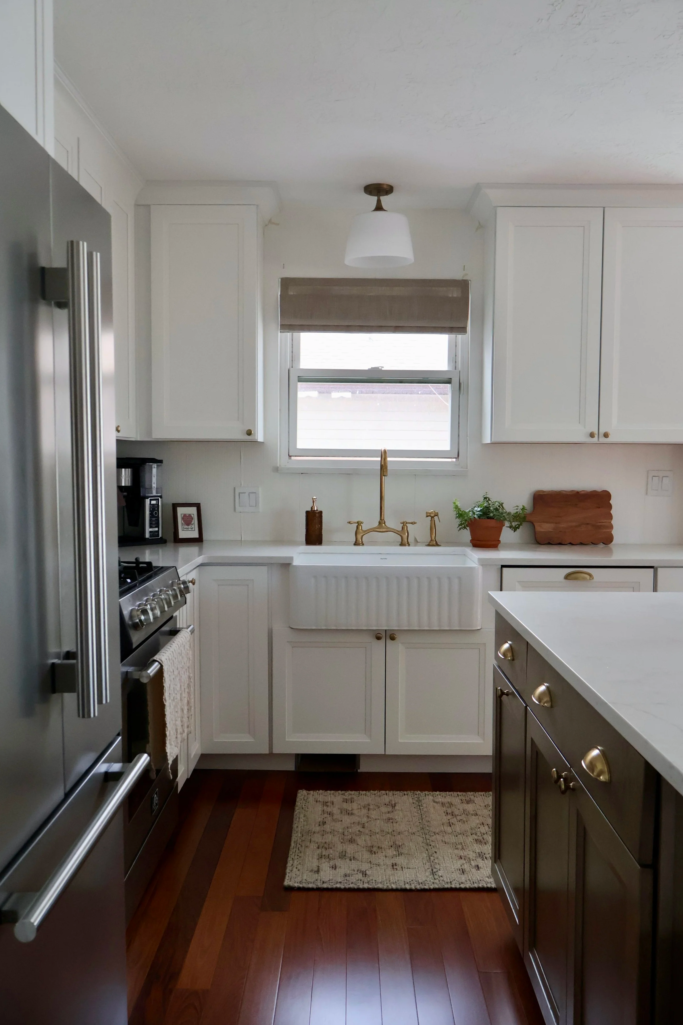 Kitchen with white cabinets, a window behind the sink, a brass faucet, a gray refrigerator on the left, a stove with an oven, a coffee maker, small framed picture, and a beige rug on brown hardwood floors.