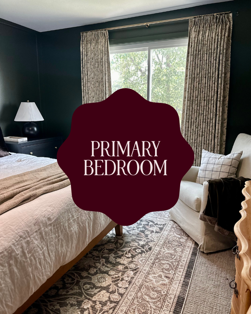 A cozy primary bedroom with a bed covered in beige bedding, a black nightstand with a white lamp, and a window with floral curtains revealing greenery outside. There is also a white armchair with a plaid pillow and a black blanket, and a patterned rug on the floor.