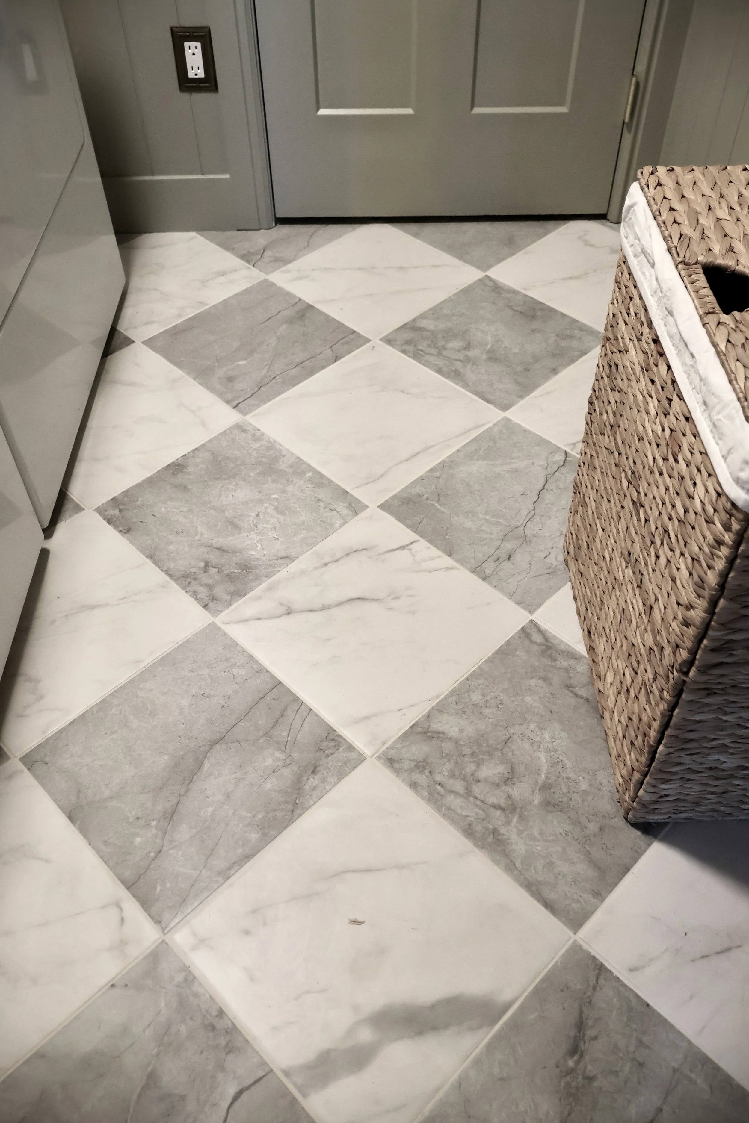 A tiled bathroom or laundry room floor with a pattern of alternating gray and white marble tiles, a gray door, an electrical outlet, a wicker basket on the right side, and part of a white appliance on the left side.