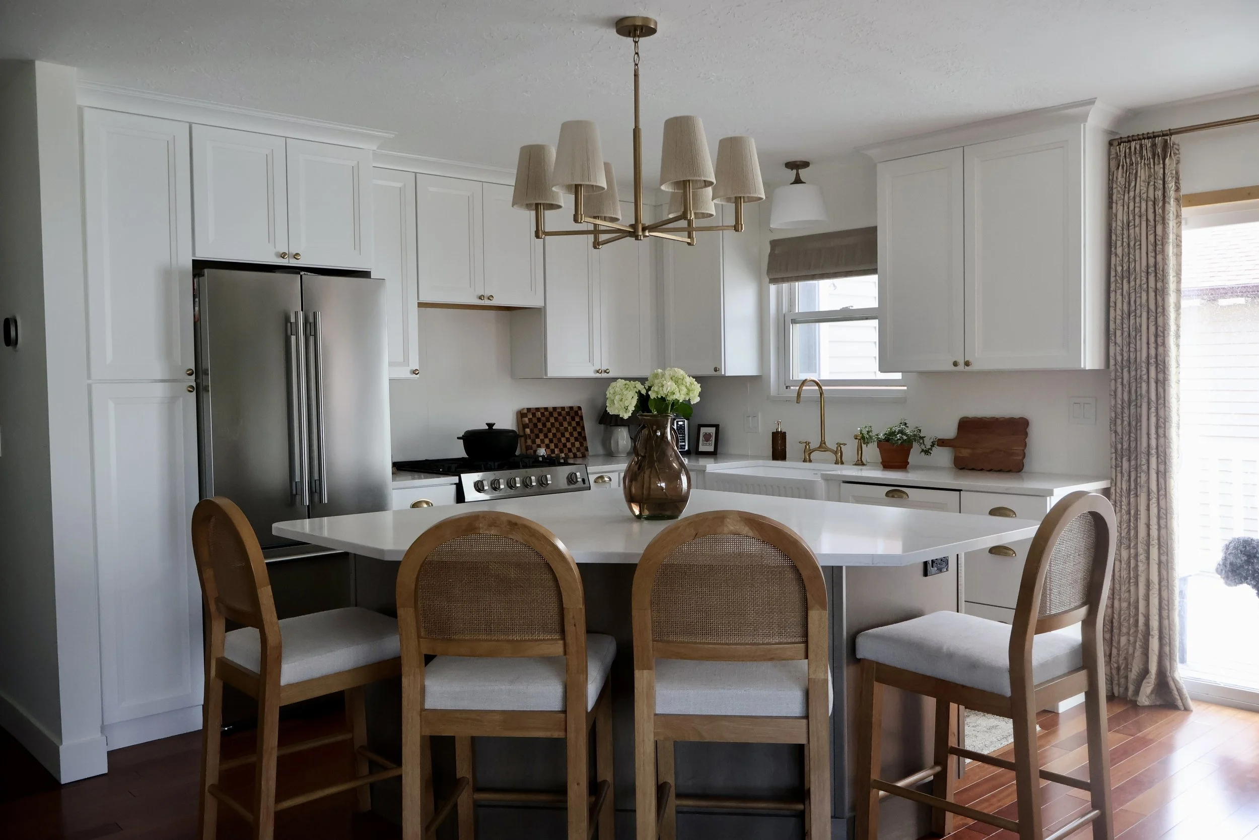 A bright kitchen with white cabinets, a large kitchen island with seating, a stainless steel refrigerator, a gold chandelier, and a window with curtains.