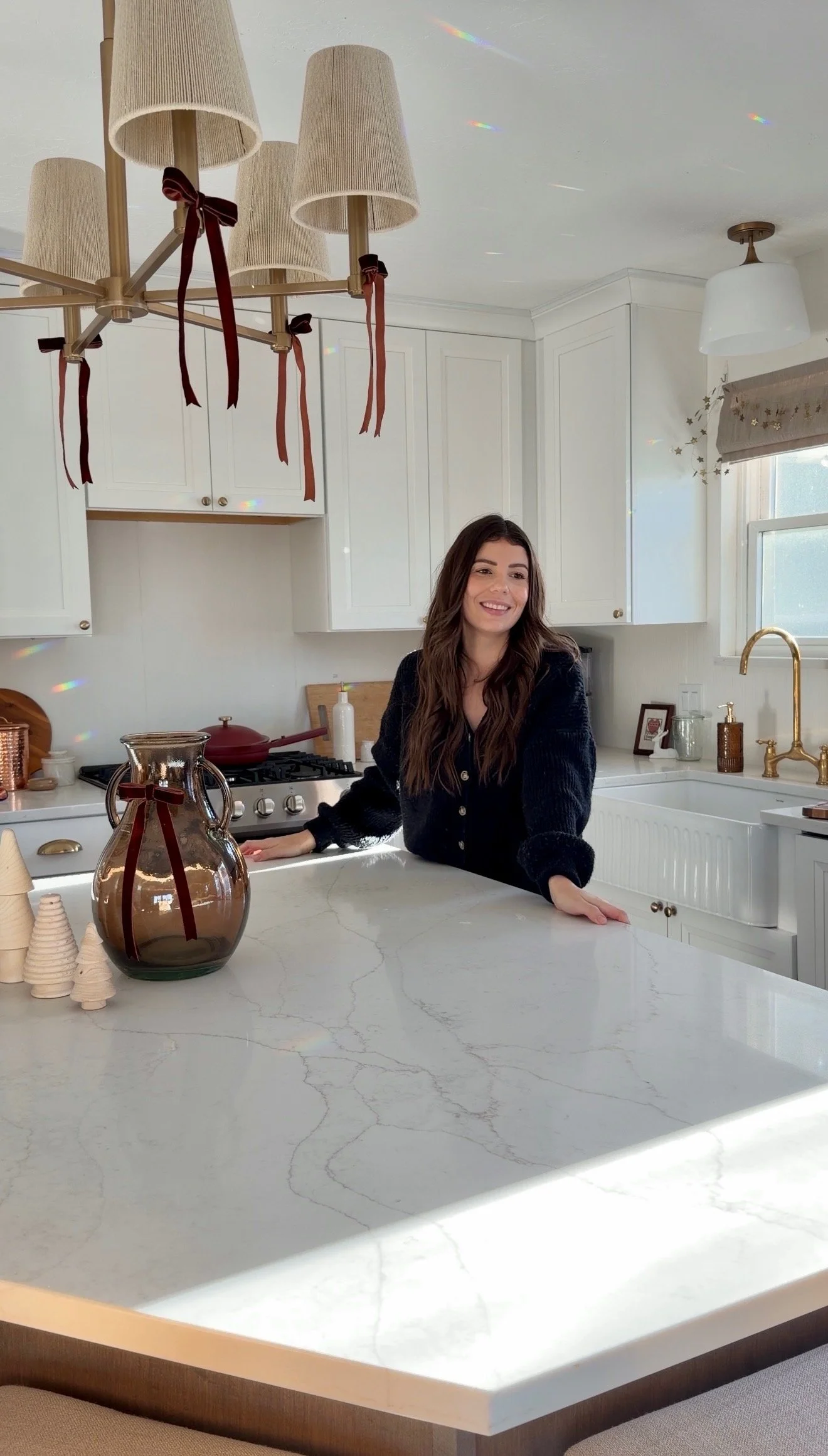 A woman with long dark hair smiling and leaning on a marble kitchen counter in a bright, modern white kitchen with gold accents and decorative items.