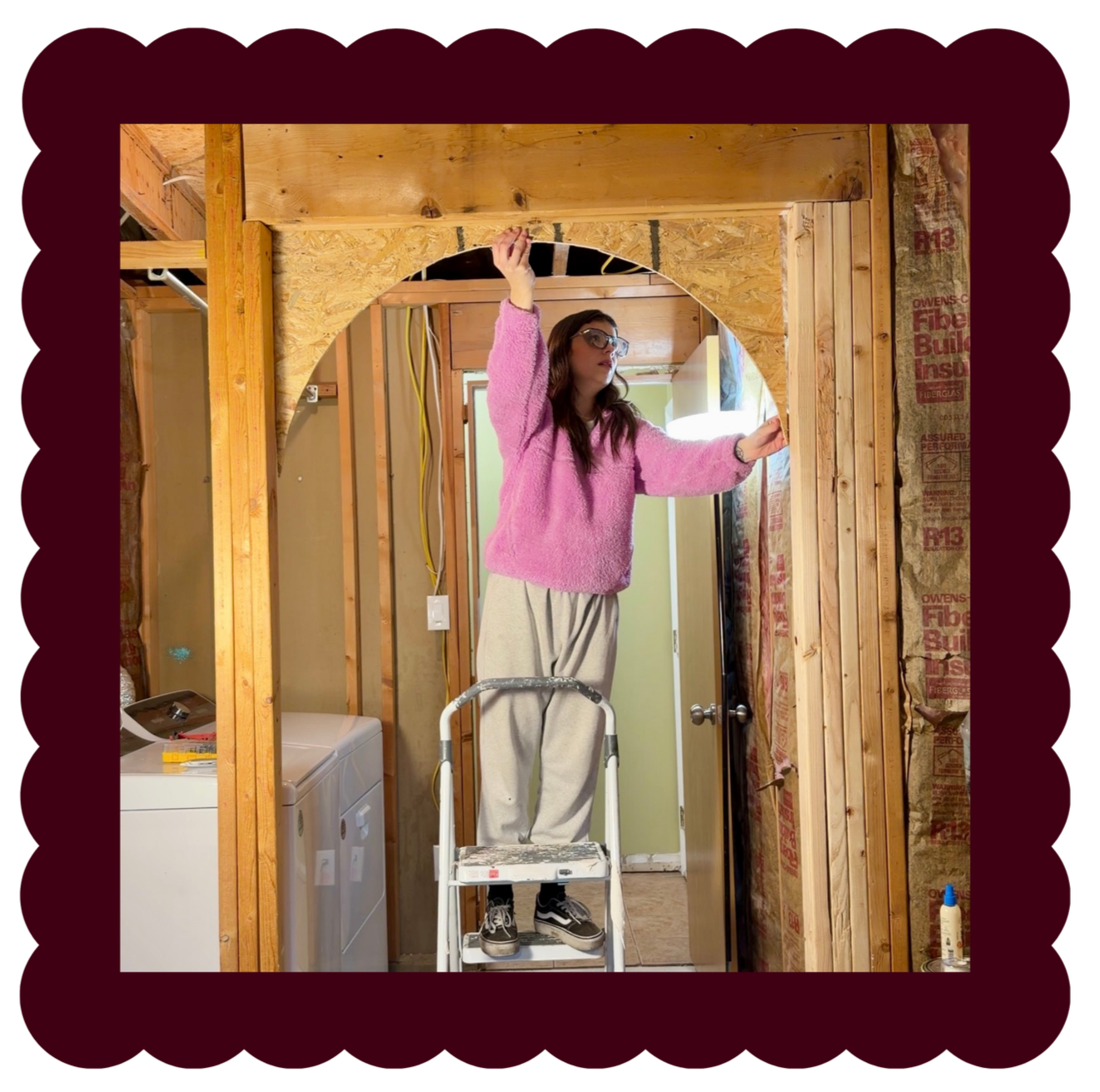 Kristen Ontiveros a woman DIYer standing on a step stool working on framing a doorway in an unfinished room with exposed wooden studs and insulation, wearing a pink sweater and glasses.