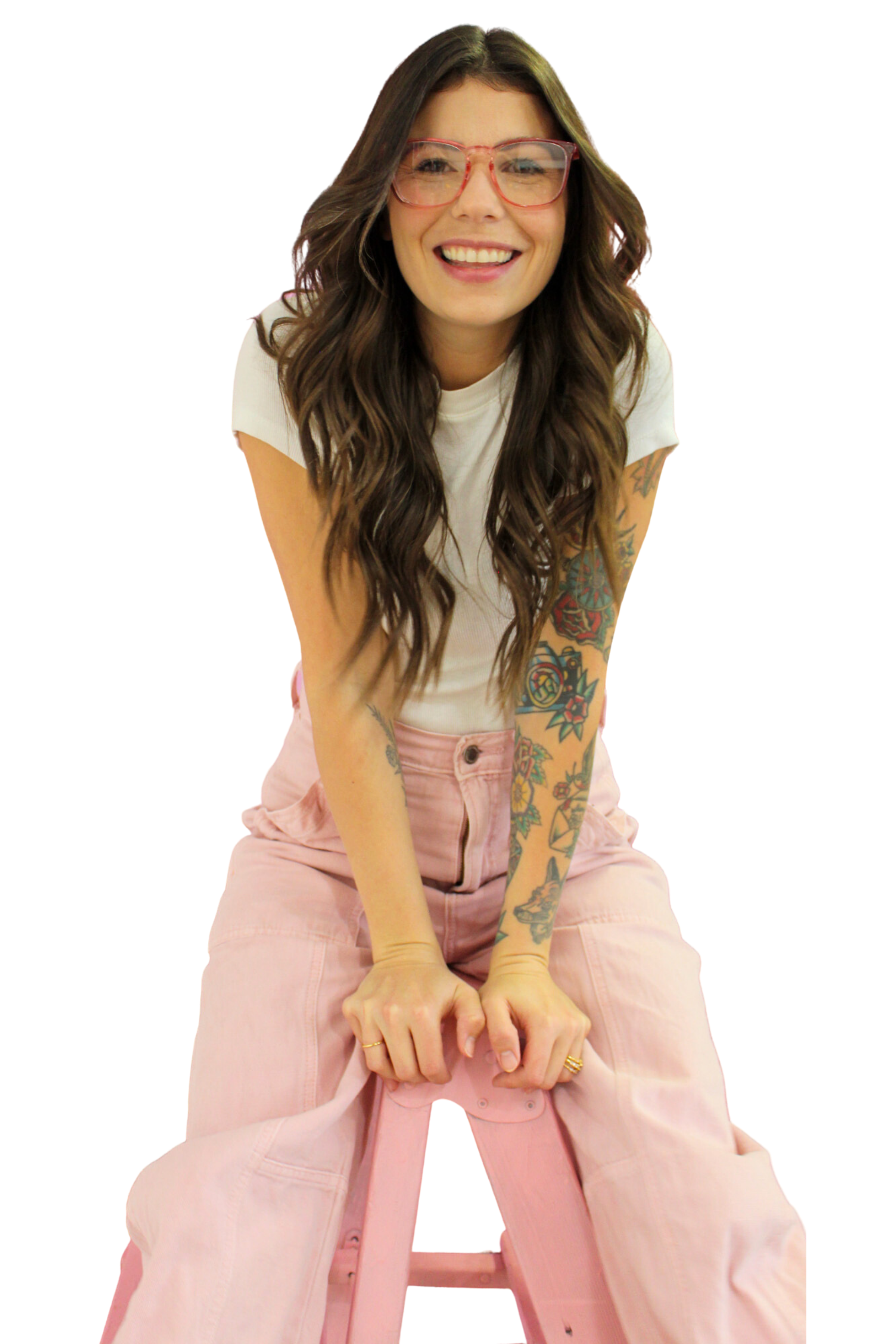 A young woman with long wavy brown hair and glasses smiling, sitting with her hands between her legs, wearing a white t-shirt and pink pants, with tattoos on her arms.