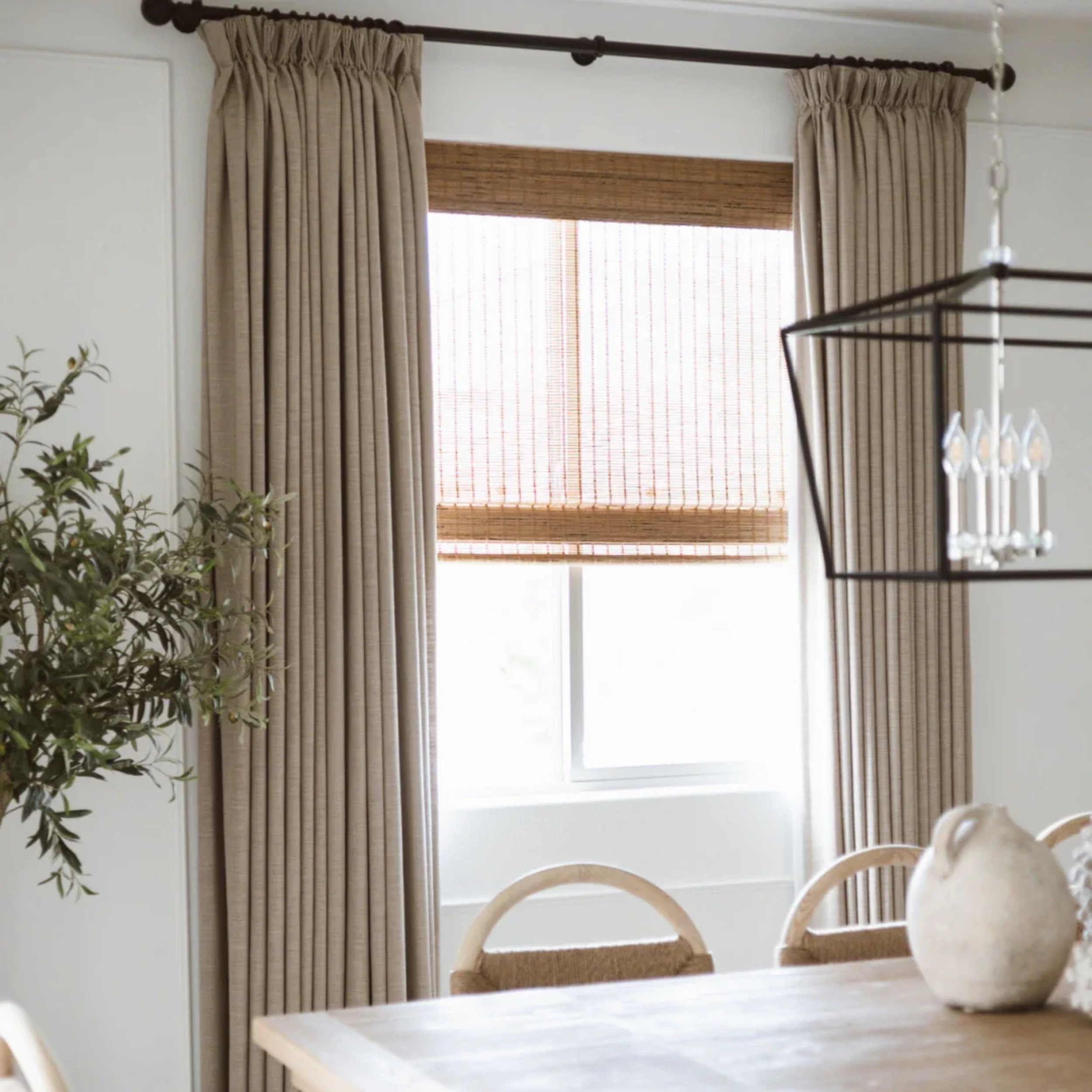 A corner of a bright room with beige curtains hanging on a dark brown curtain rod, covering a window with brown woven blinds. Part of a white dining table and chairs with light-colored upholstery are visible. A leafy plant is in the lower left corner.