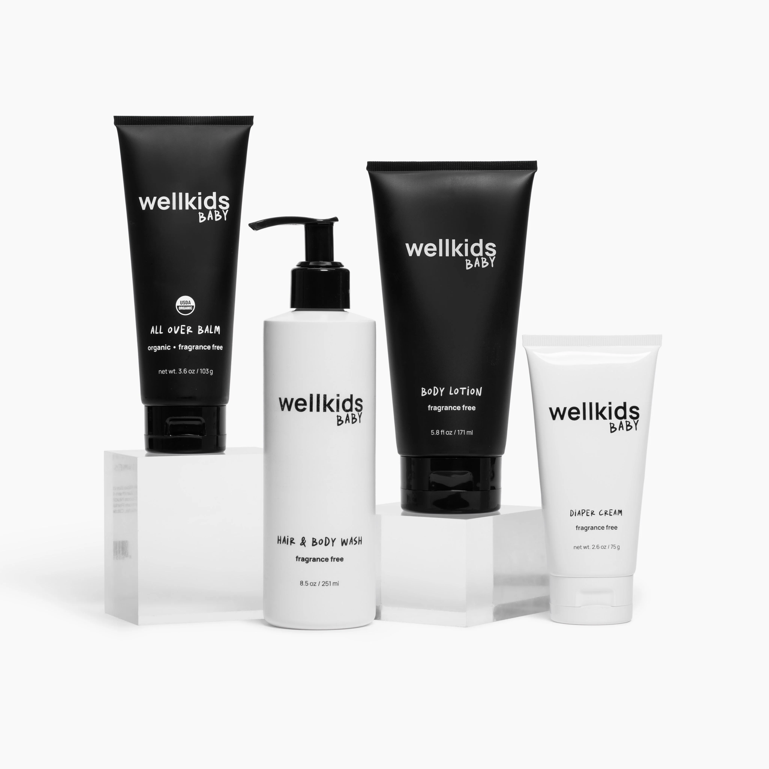 Collection of Wellkids Baby skincare products including all-over balm, hair & body wash, body lotion, and diaper cream, displayed on white stands with minimalist black and white packaging.