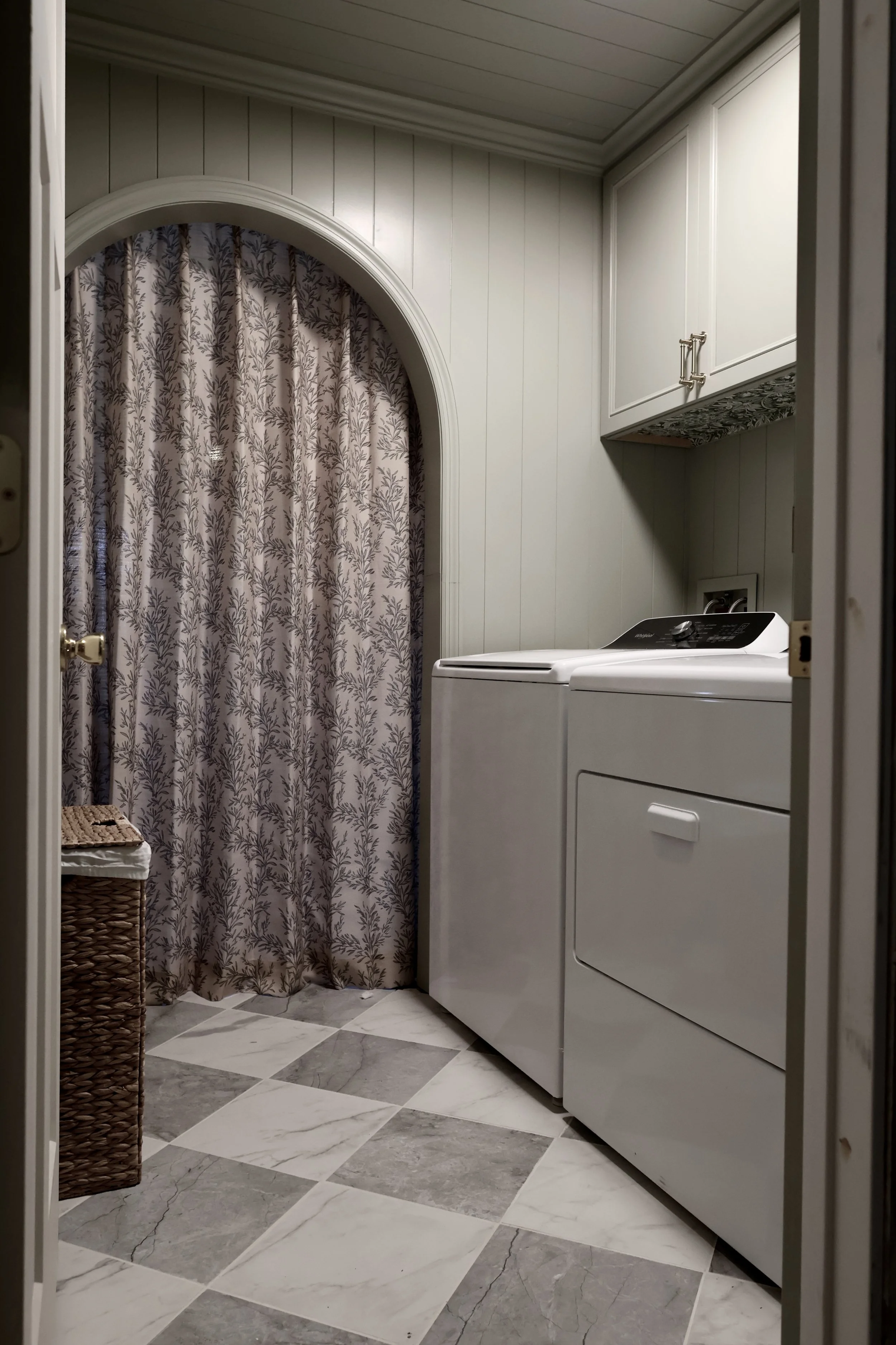 Laundry room with a white washing machine and dryer, white cabinets above, a patterned curtain, a basket, and marble tile flooring.