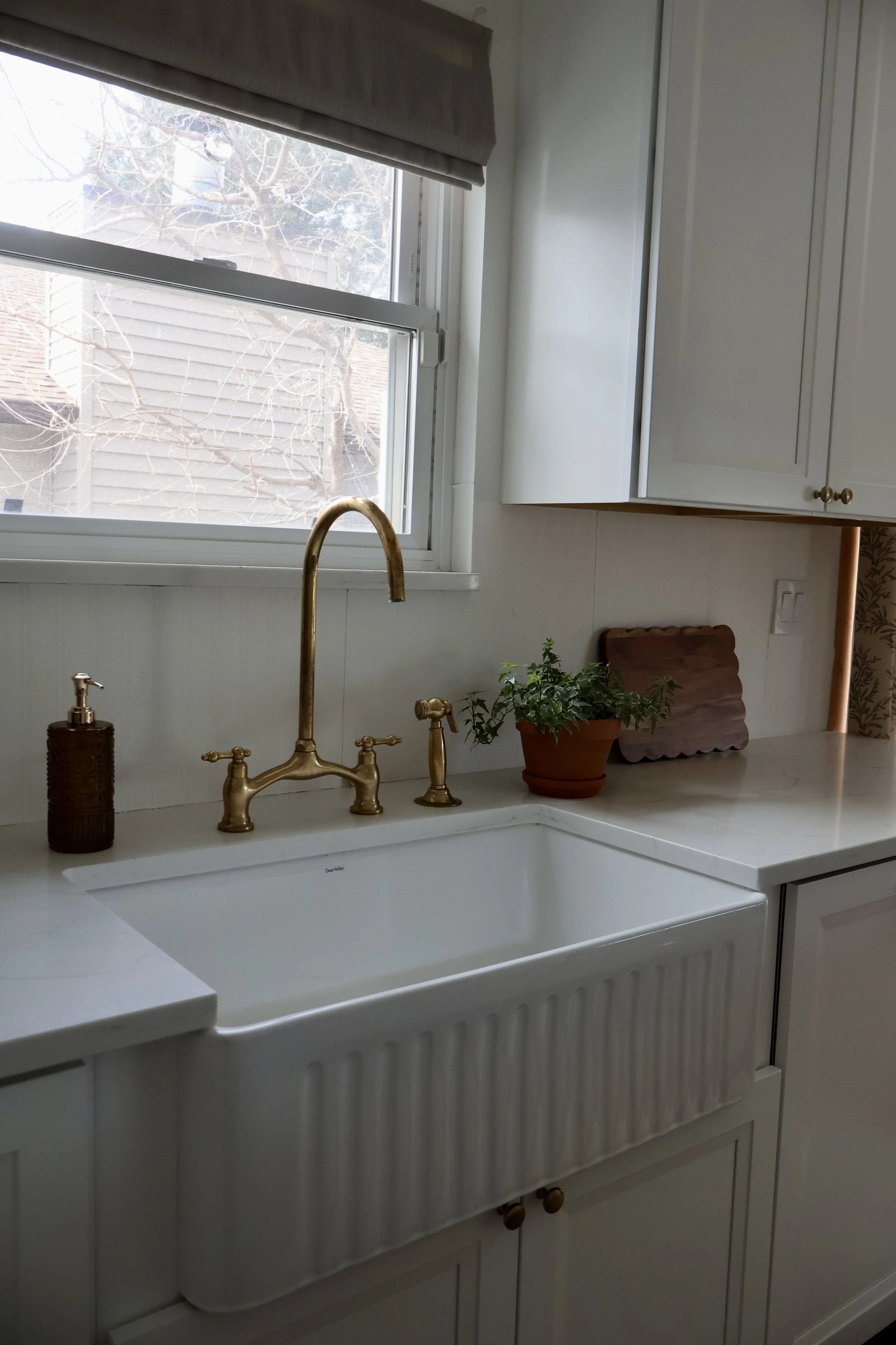 Kitchen sink with gold-colored faucet, white cabinetry, a window with a view of trees, potted plant, soap dispenser, and a cutting board.