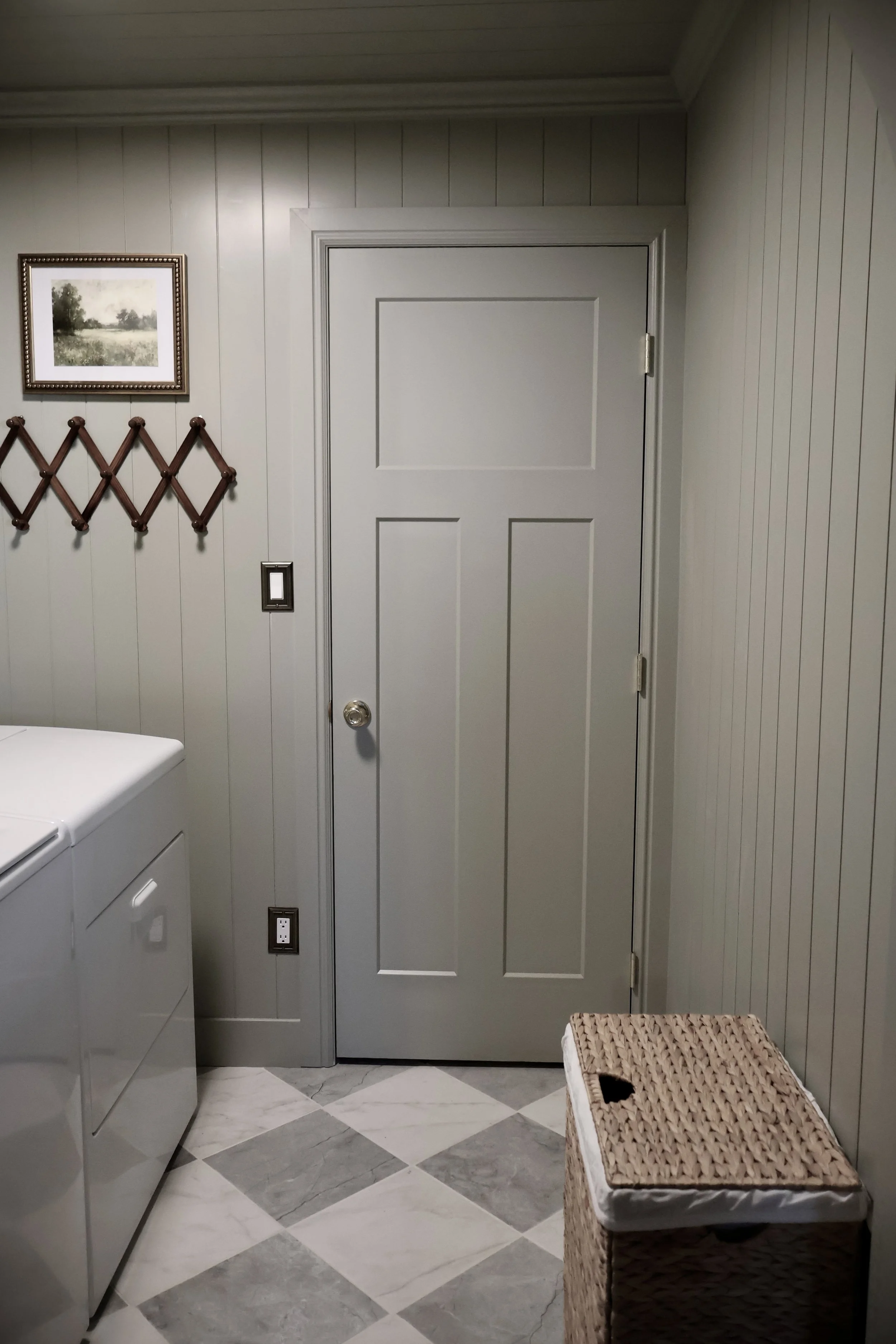 Inside laundry room with white washing machine, beige and gray tiled floor, paneled walls, framed landscape painting, wall-mounted coat rack, wicker basket, light switch, and electrical outlets.