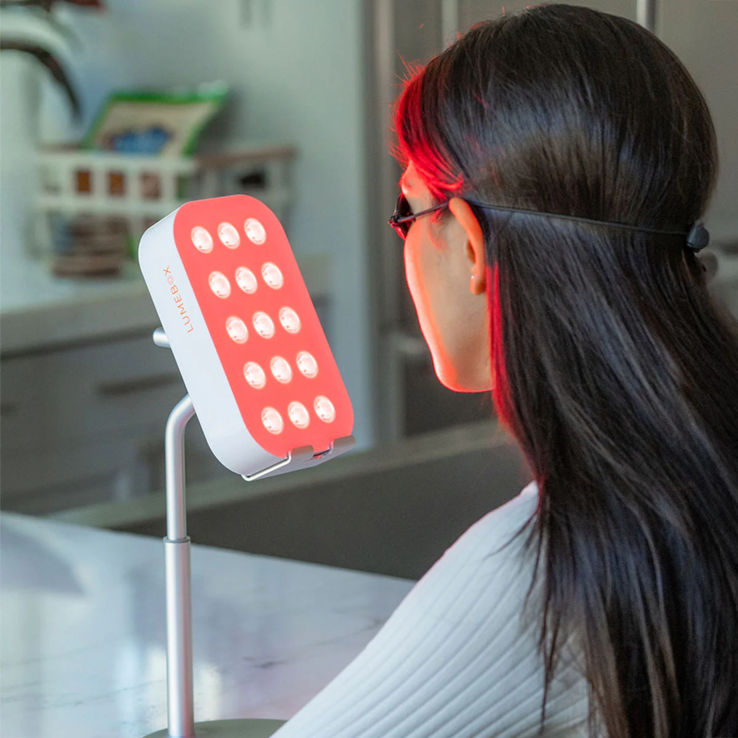 A person with dark hair and glasses is sitting in a waiting area, facing away, with a pink medical lamp nearby and a blurred background.