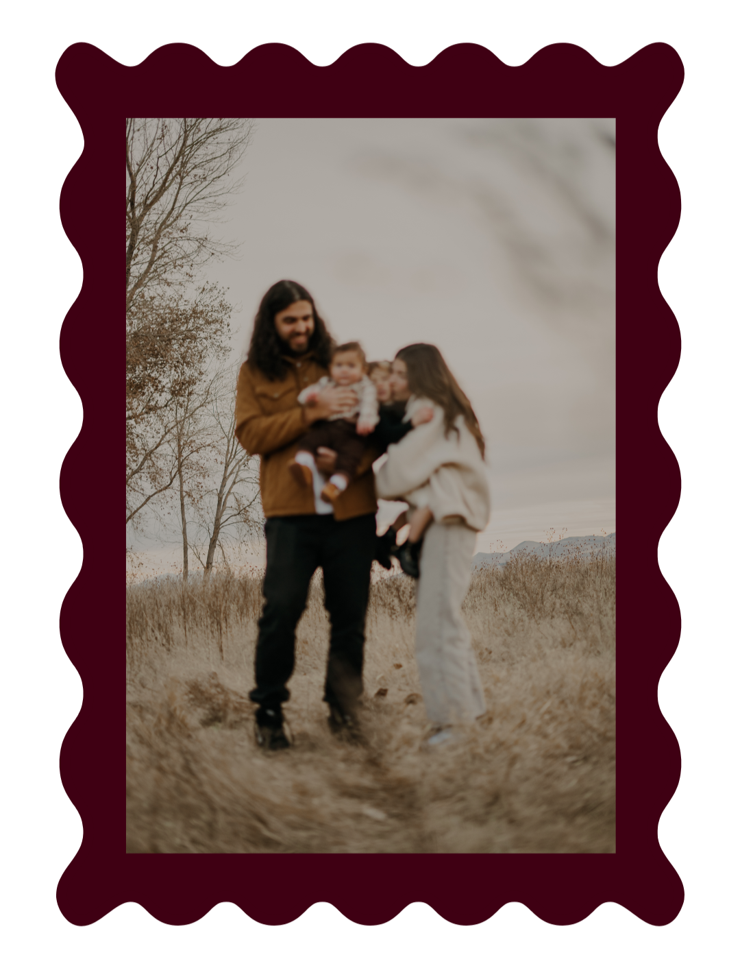 A family of four standing outdoors in a field during autumn, smiling and embracing each other. The man has long hair and a beard, holding a baby, while a woman and a young girl stand close beside him.