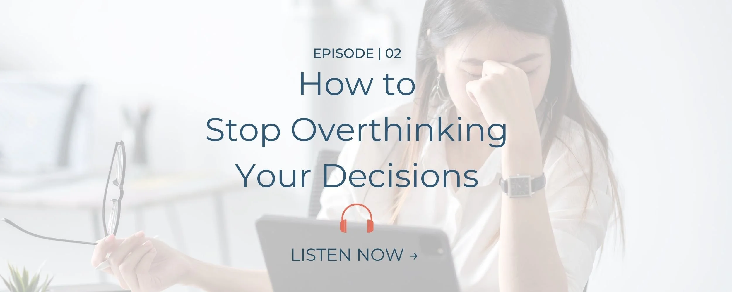 Asian woman sitting at desk holding reading glasses with her head in her hands, looking stressed.