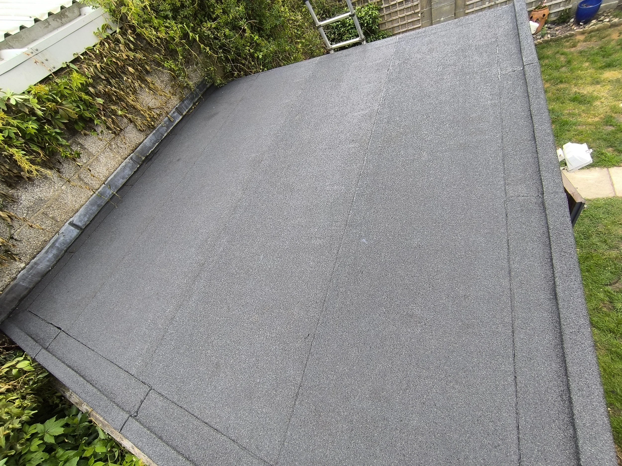 Flat roof covered in black roofing felt with a slight ridge in the middle, bordered by greenery and a brick wall with ivy on the left and a garden with grass and paving stones on the right.