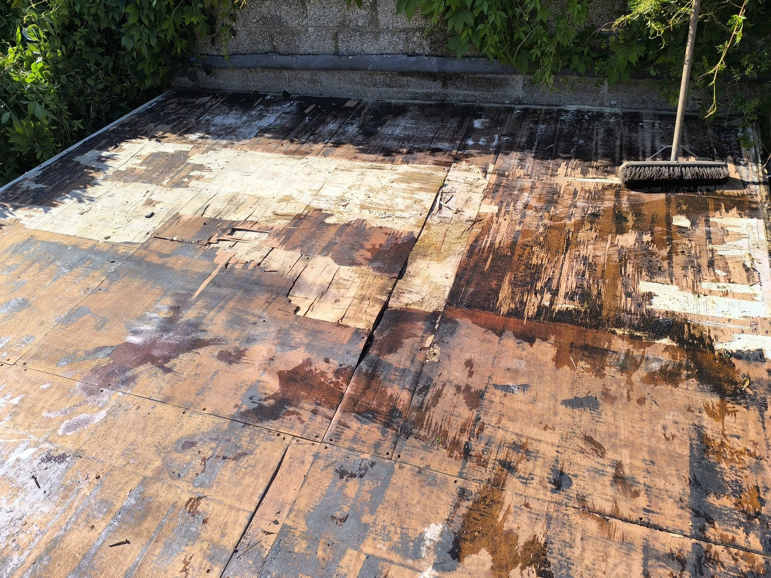 Weathered and peeling wooden roof with a broom leaning against the edge, surrounded by green foliage.