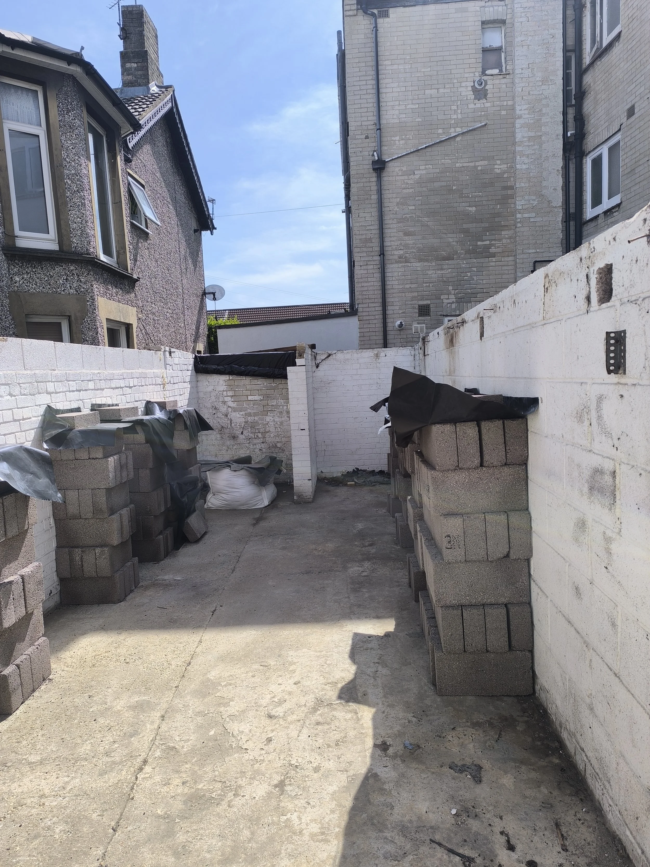 Construction site with stacked concrete blocks along white brick wall, bags of materials, and a clear blue sky.
