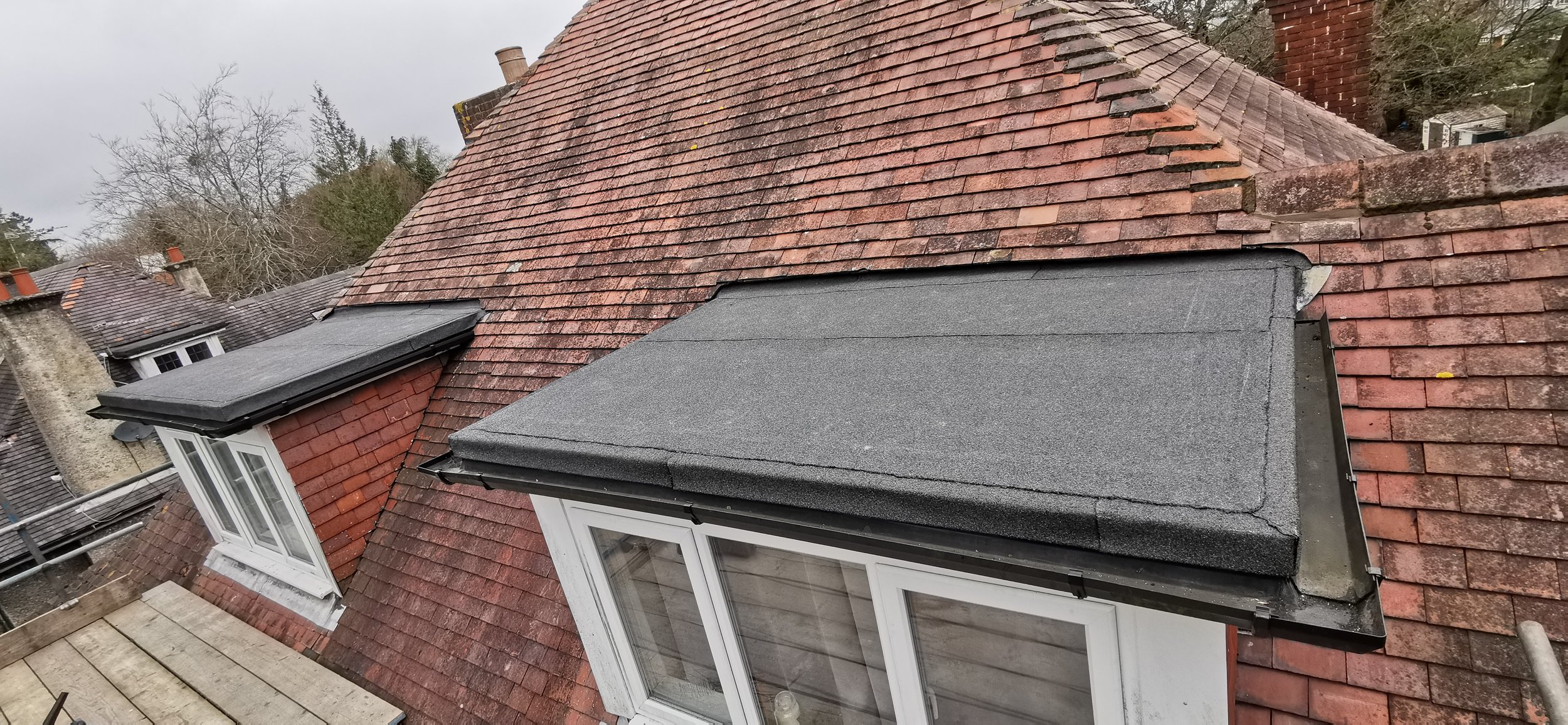 Two sections of flat roofing with black and gray materials on top of white-framed windows on a brick house, surrounded by a red-tiled sloped roof.