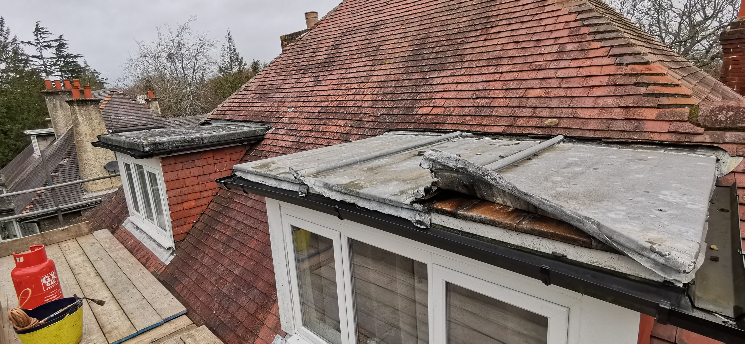 Views of a roof with old and damaged roofing material, including construction supplies and equipment such as a propane tank on a wooden scaffolding platform.