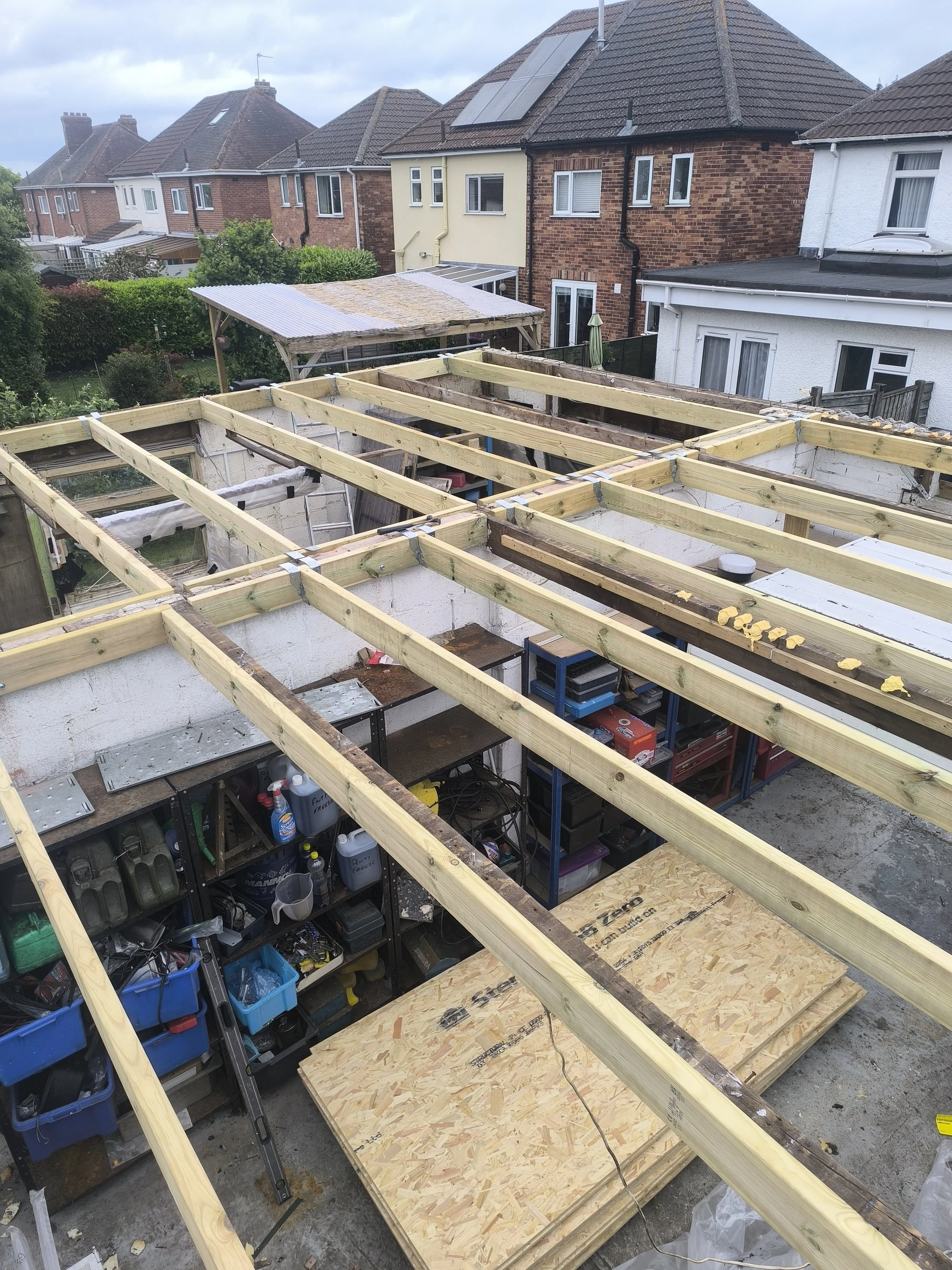 Construction site with wooden beams being installed on top of a building, neighboring houses in the background, and shelves with tools underneath.