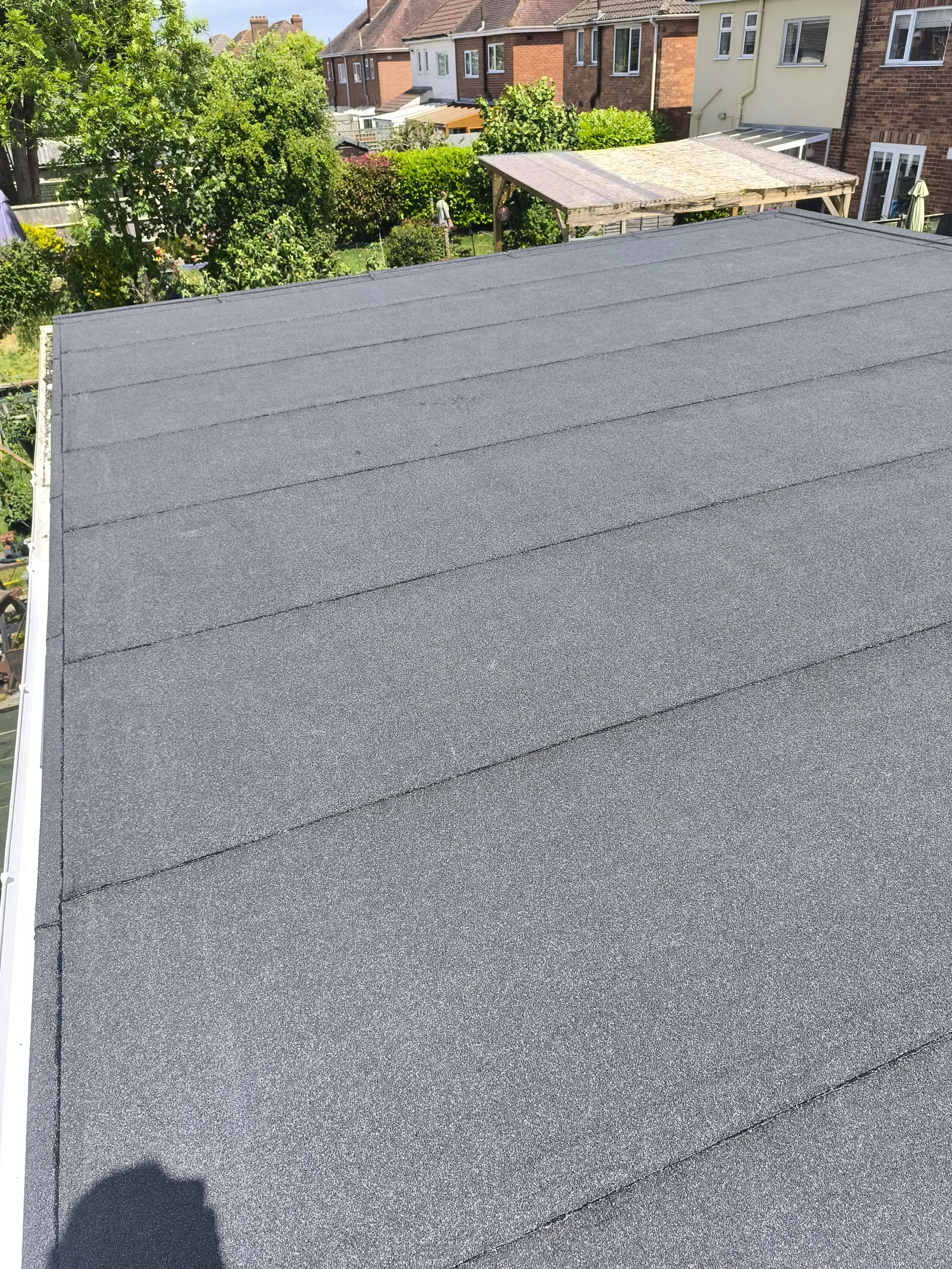 Flat rooftop covered with new black asphalt shingles, with neighboring houses and greenery in the background under a clear blue sky.