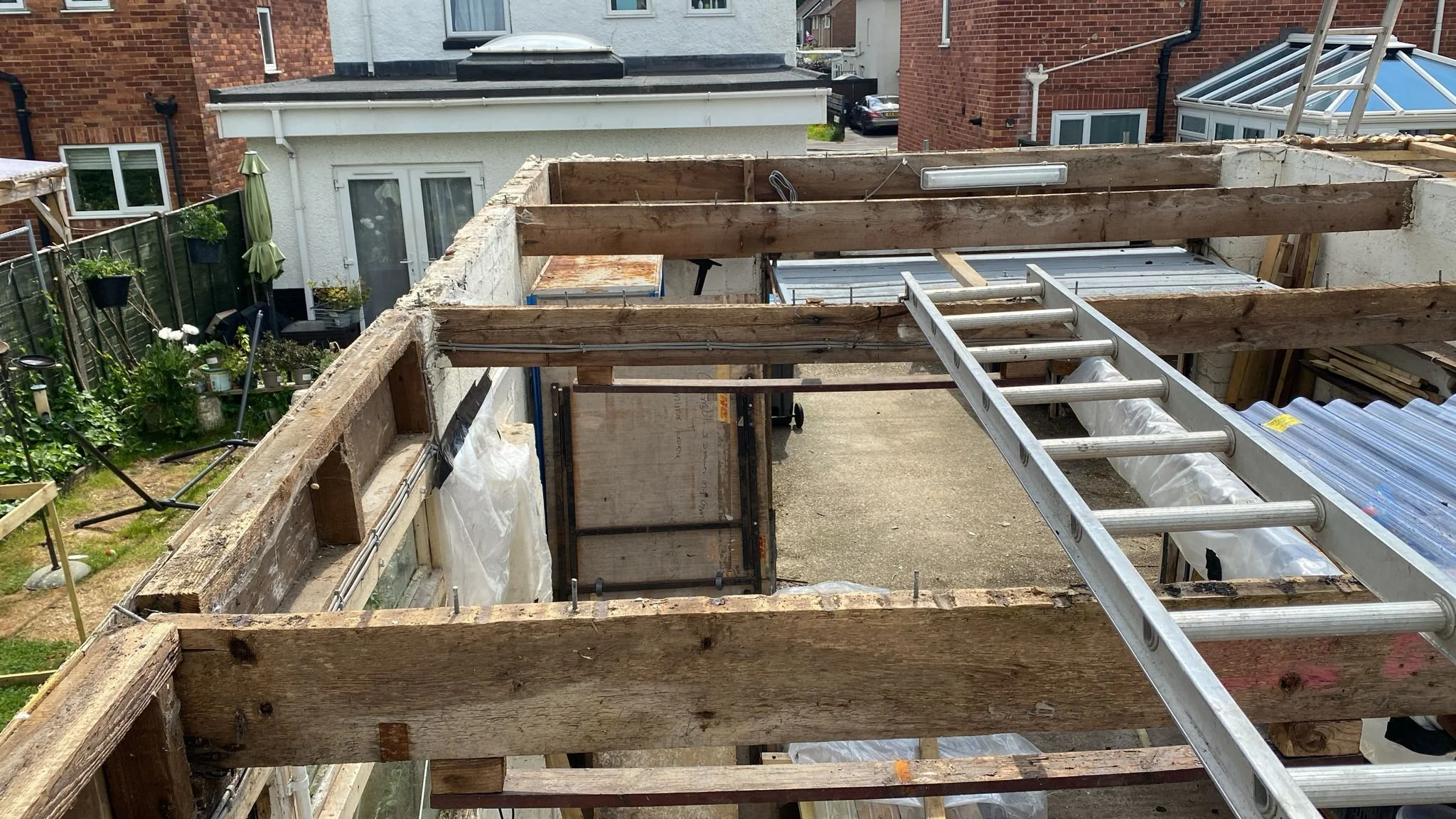 View of a construction site on a building with exposed wooden beams and a ladder, overlooking neighboring houses and a backyard garden with plants and outdoor furniture.