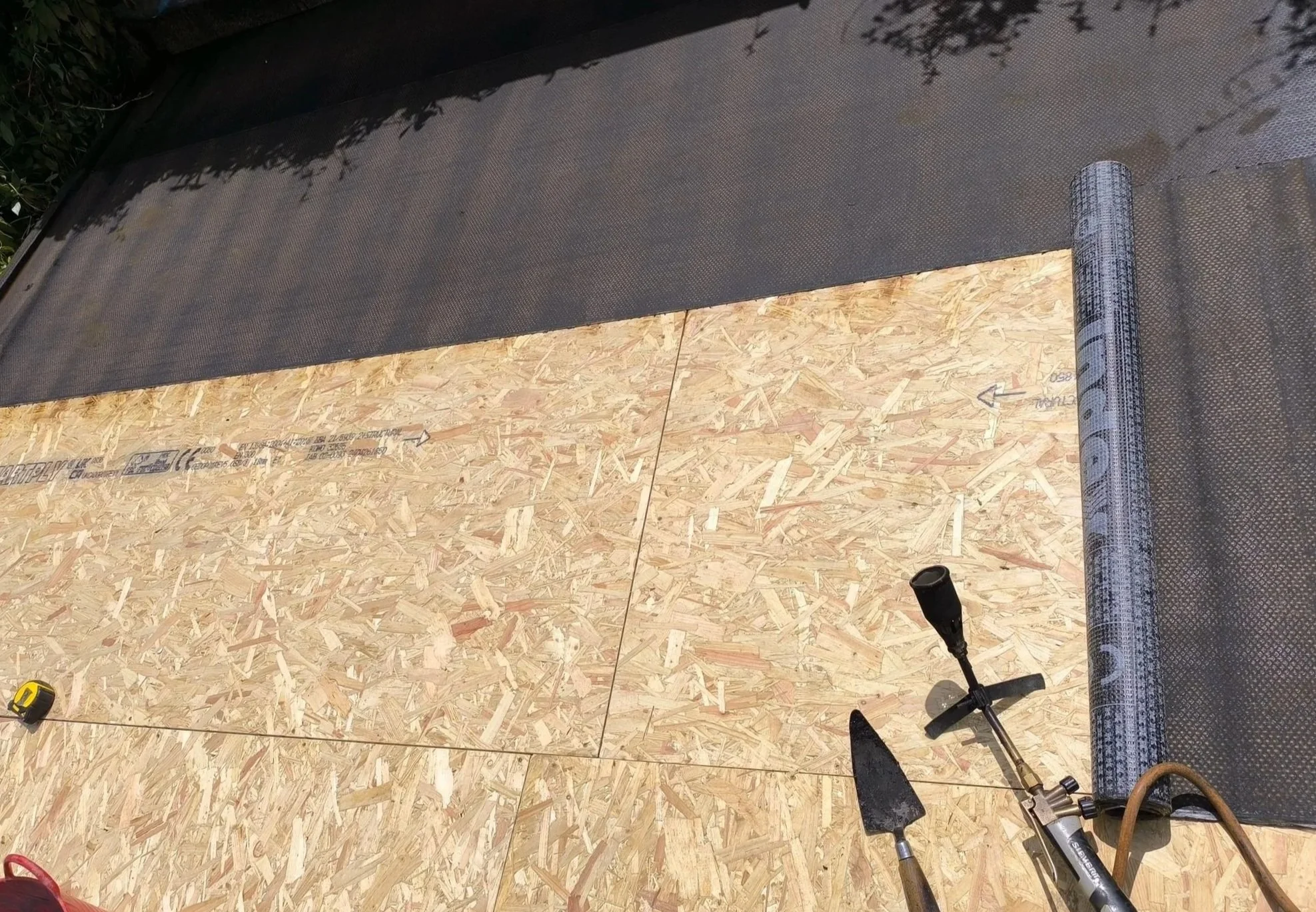 OSB plywood sheet being installed on a roof, with tools nearby including a trowel and a roll of roofing underlayment.