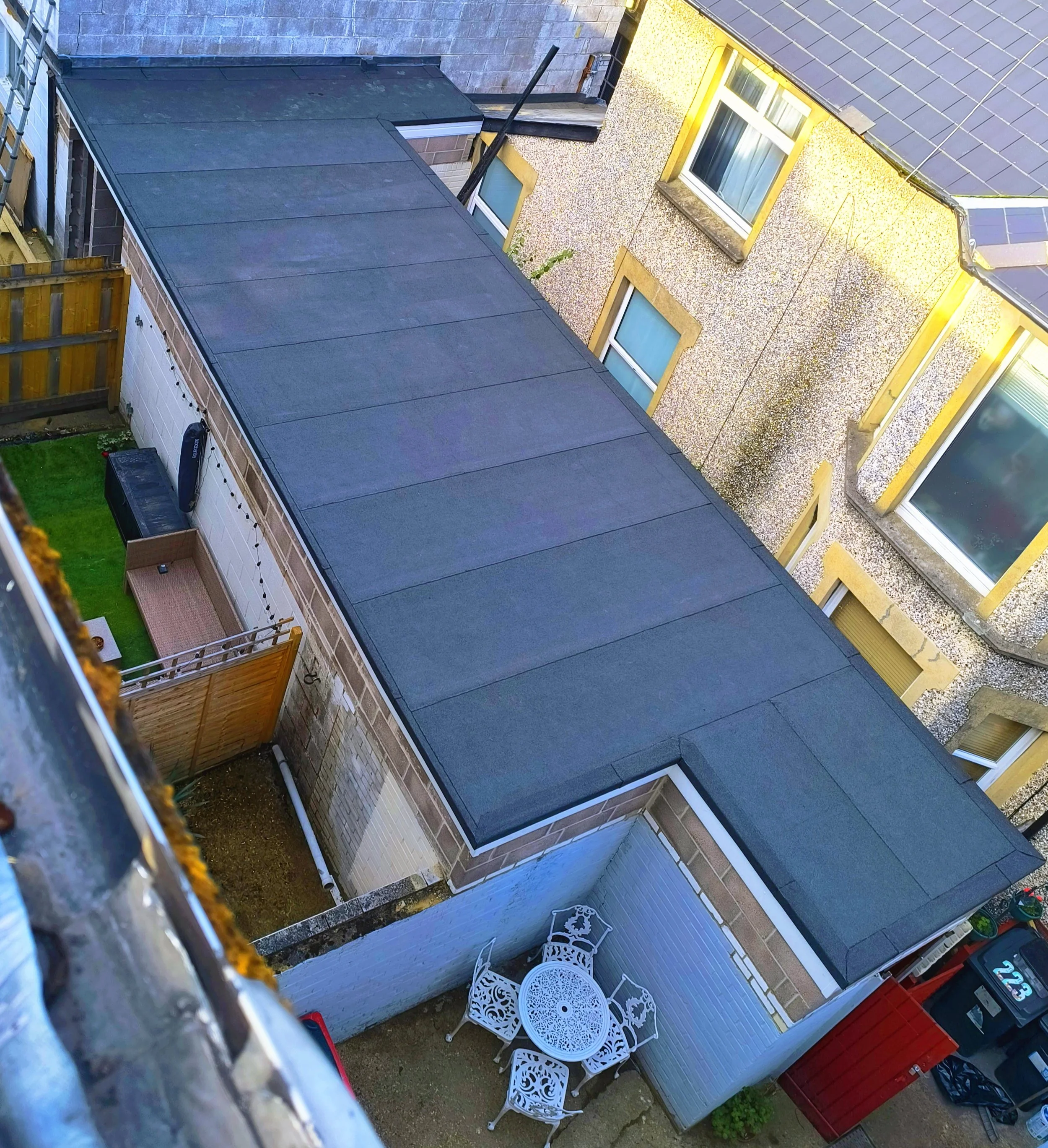 View from above of a backyard with a black flat roofed extension, a white fence with a round table and four chairs, a small patch of green grass, and a wooden gate. The backyard is enclosed by a building with a yellow and textured exterior wall with 
