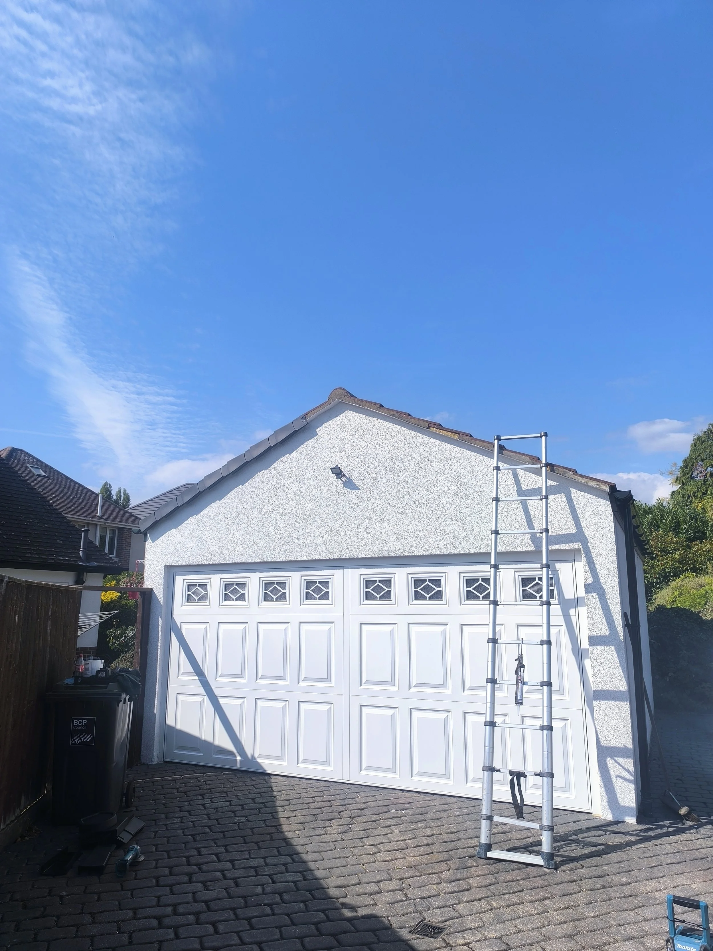 White garage with a ladder leaning against the roof, a black trash bin, and a shadow cast on a cobblestone driveway under a clear blue sky.