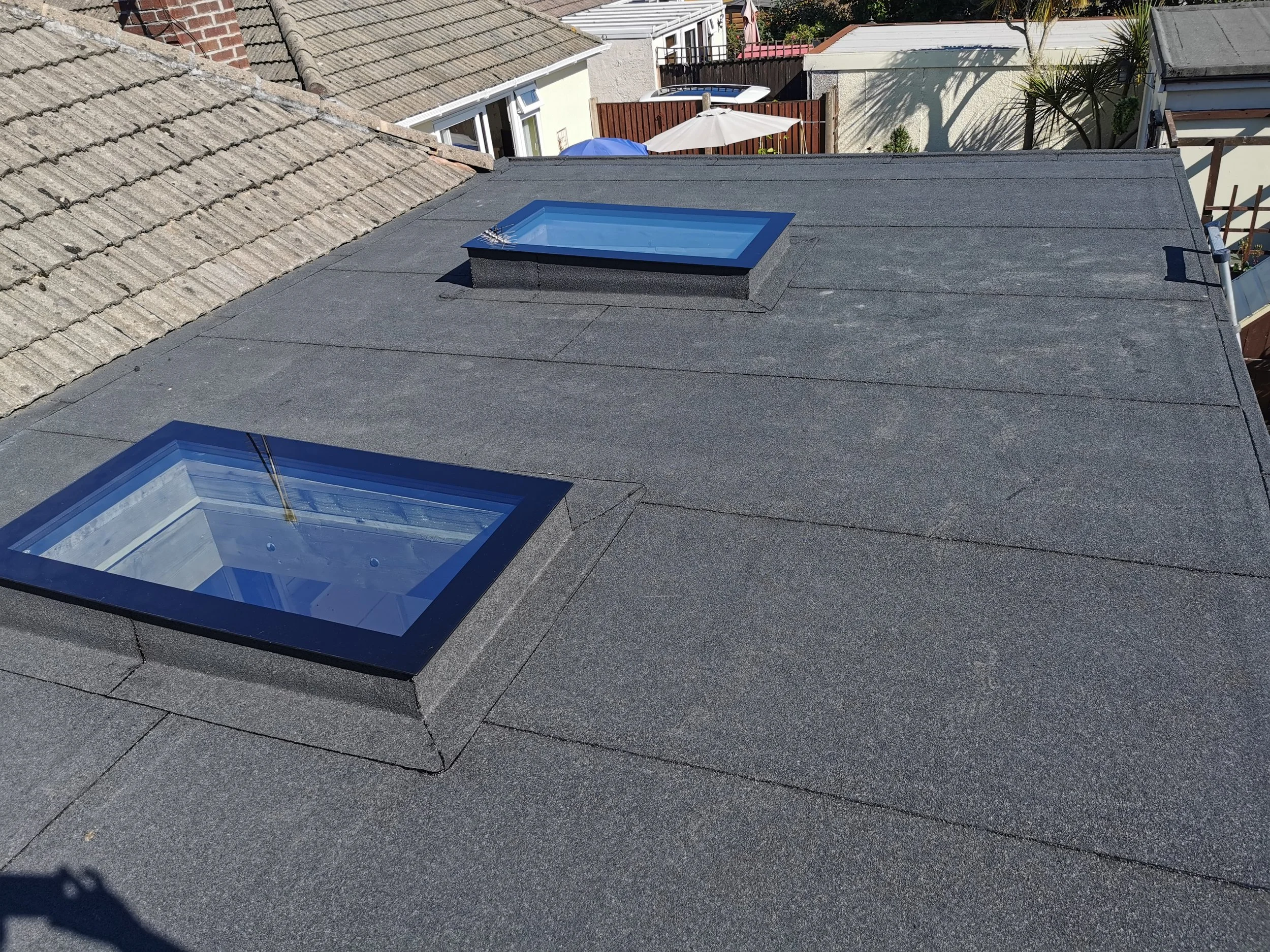 Looking down on a flat black roof with two skylights, one larger and one smaller, both with blue frames. Surrounding rooftops with different materials and a backyard with umbrellas and trees are visible in the background.