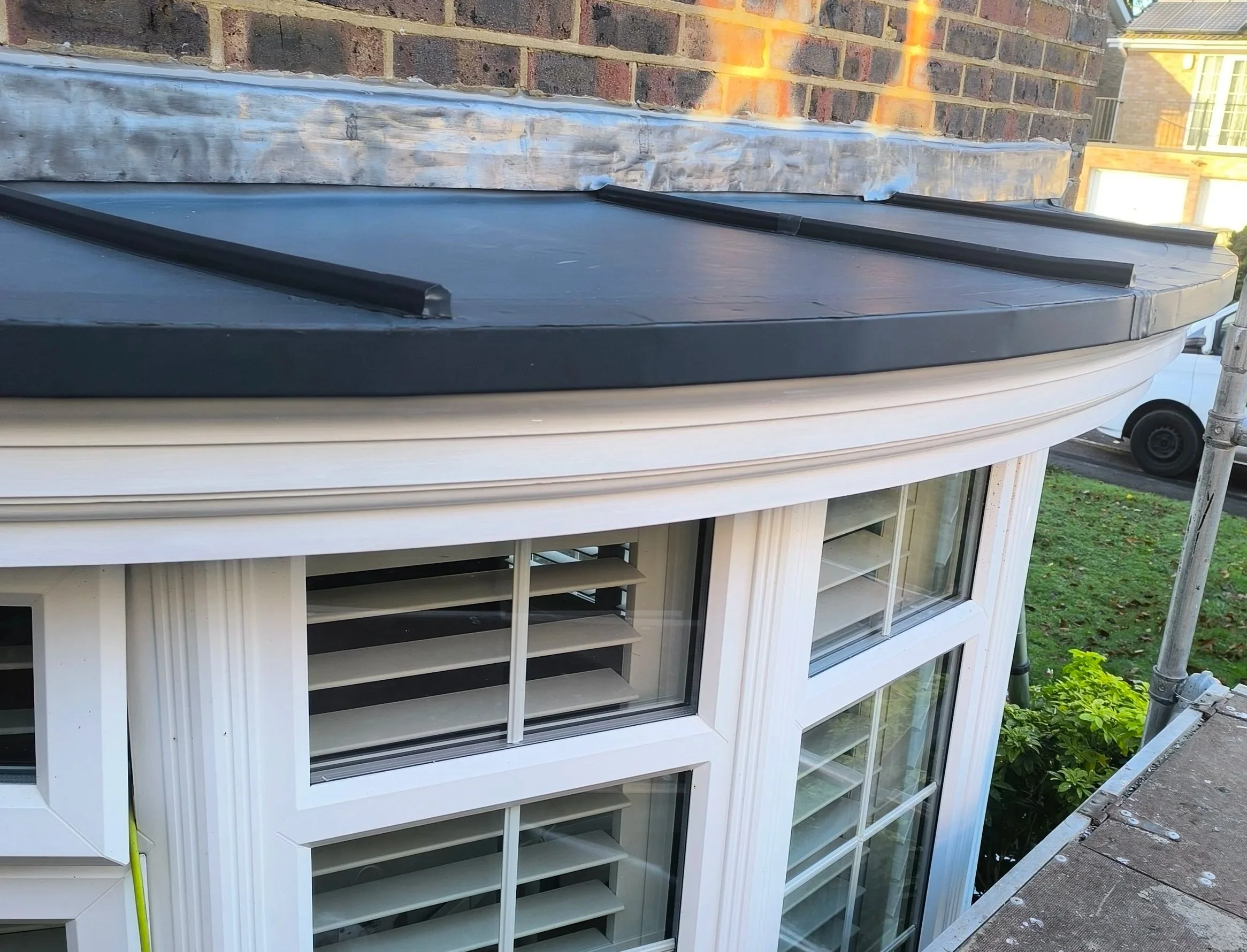 Close-up view of a building's window with white frame and adjustable security shutters, with a brick chimney and gutter in the background.
