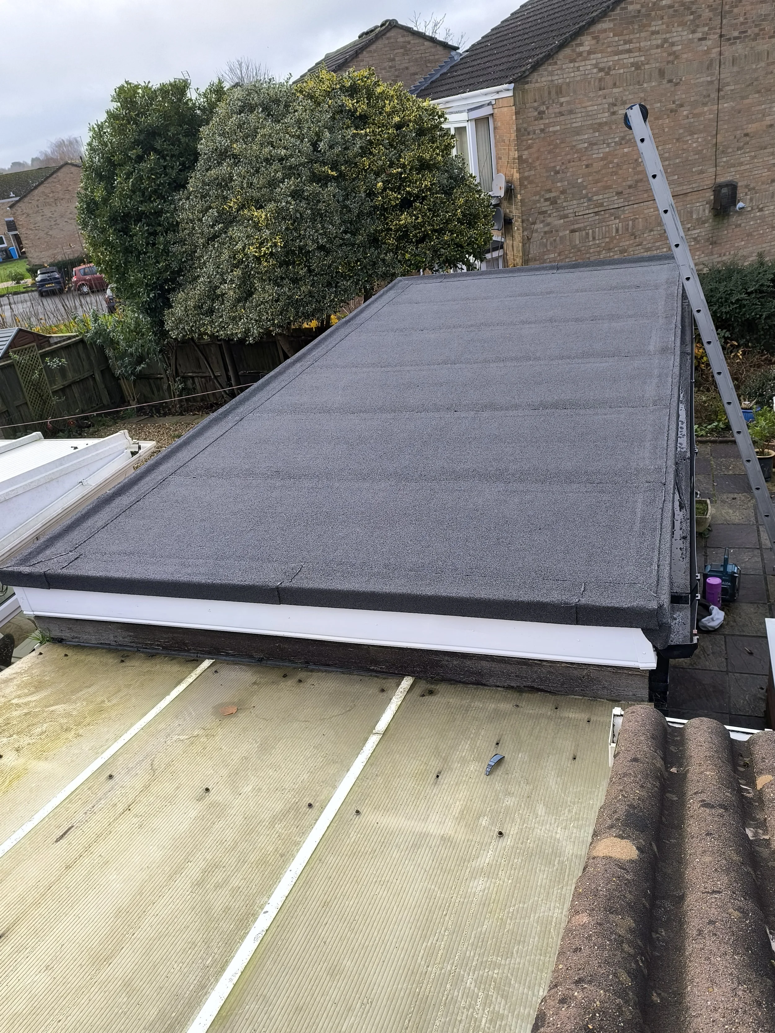 A house roof with a black felt roofing installed, with a ladder leaning against the roof's edge and various tools on the ground nearby.