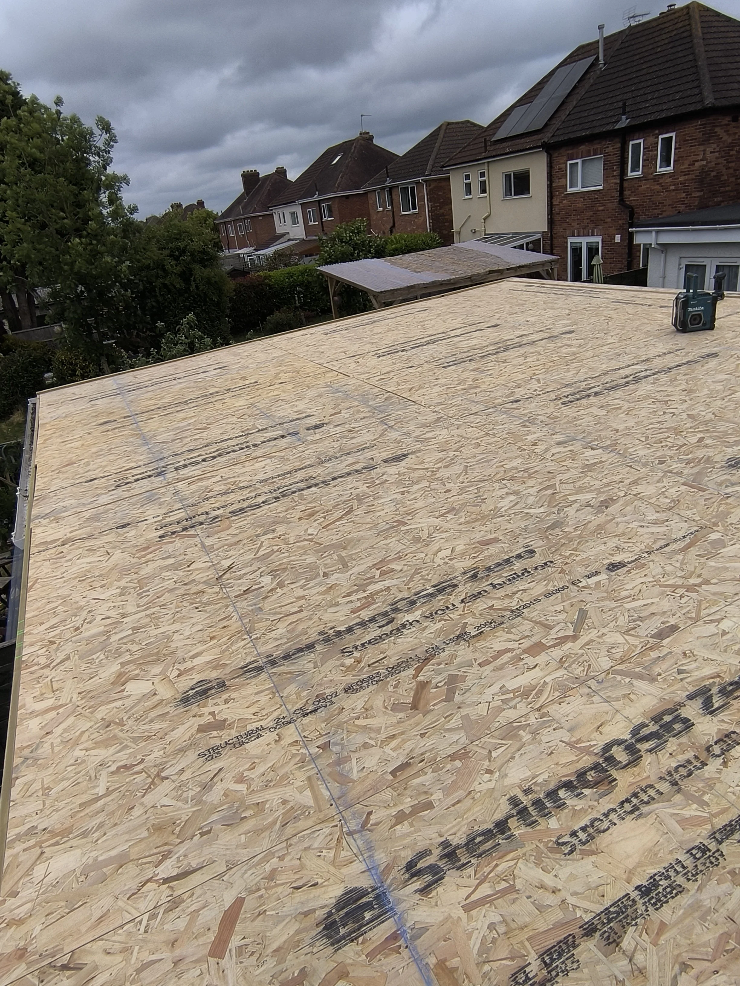 Flat roof under construction with OSB sheathing, a tape measure, and surrounding residential houses and cloudy sky.