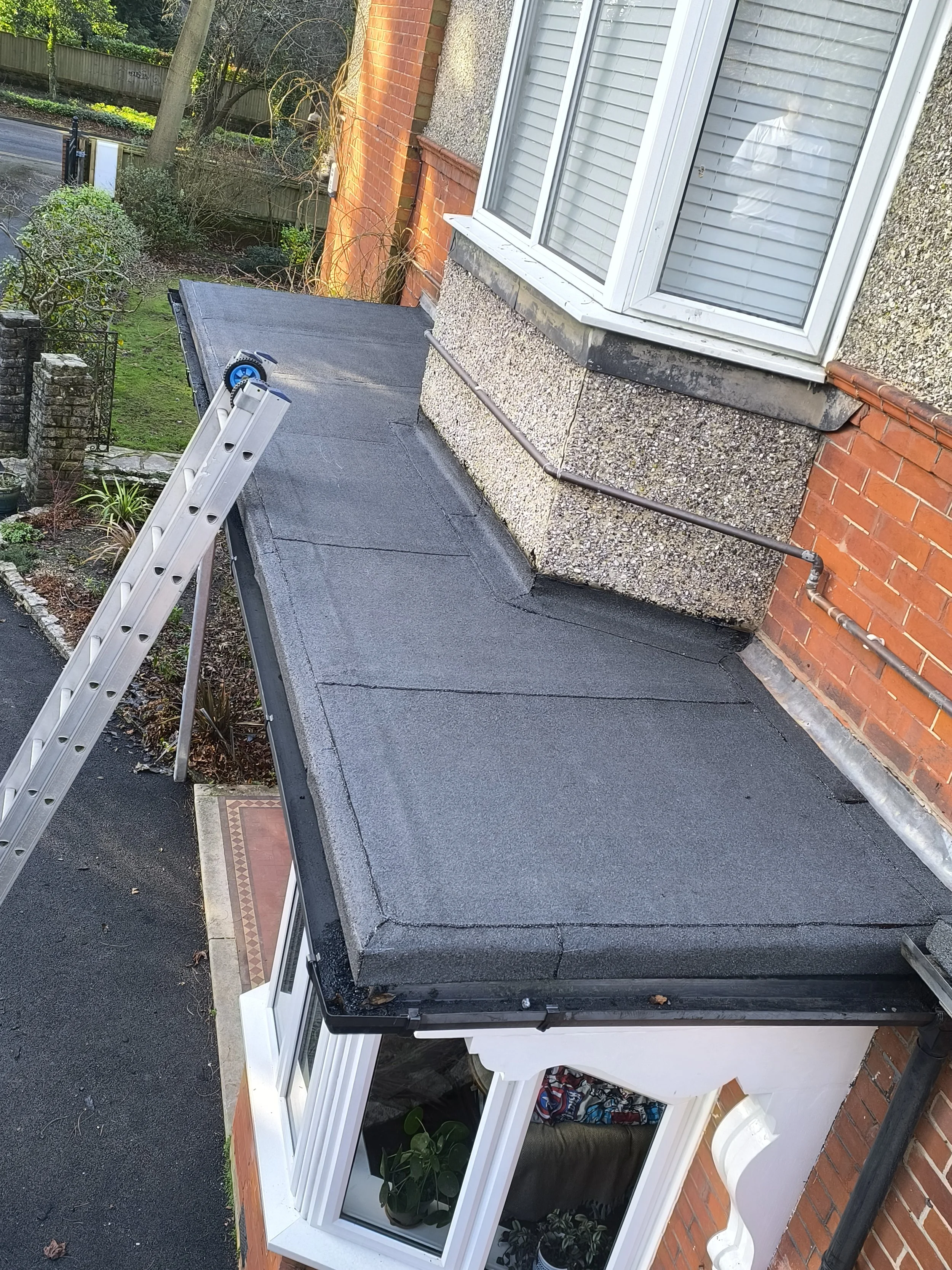 A flat roof with roofing material applied, connected to a brick wall of a house with a large window. A ladder is propped against the roof, and a person is seen in the reflection of the window taking a photo.