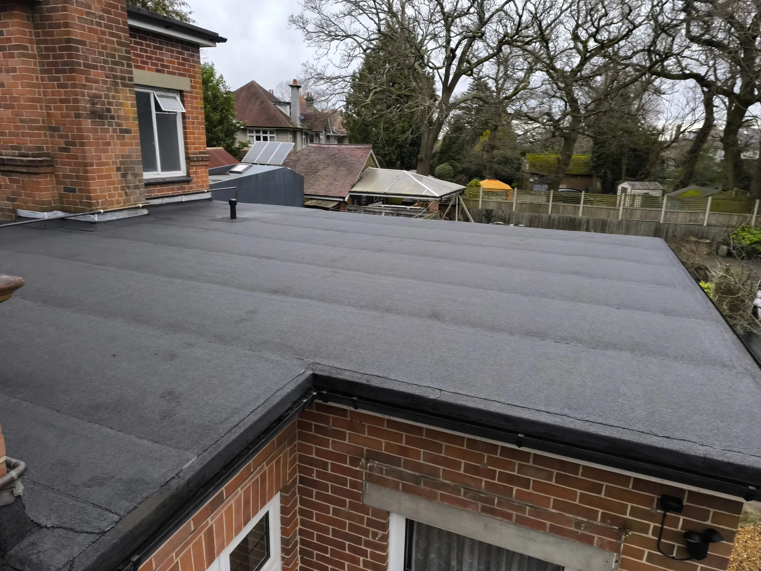 View of a flat, dark gray roof on a brick house on a cloudy day, with trees and neighboring houses in the background.