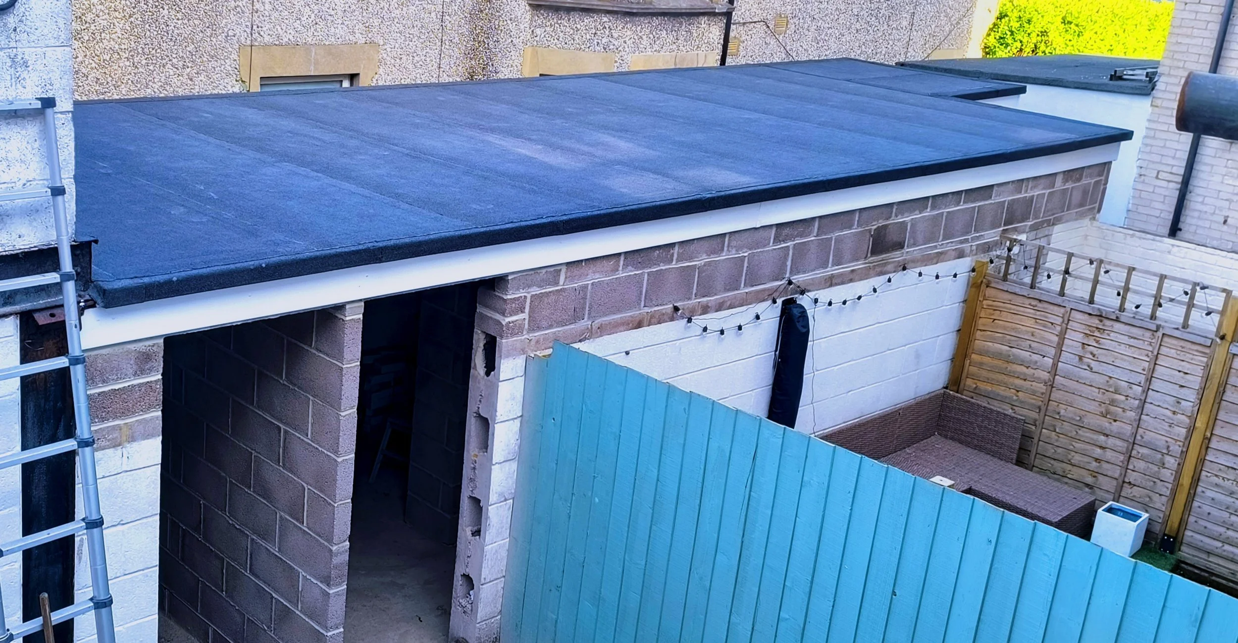 Backyard with a blue painted wooden fence, a patio with outdoor furniture, string lights, and a shed with a flat roof and brick walls.