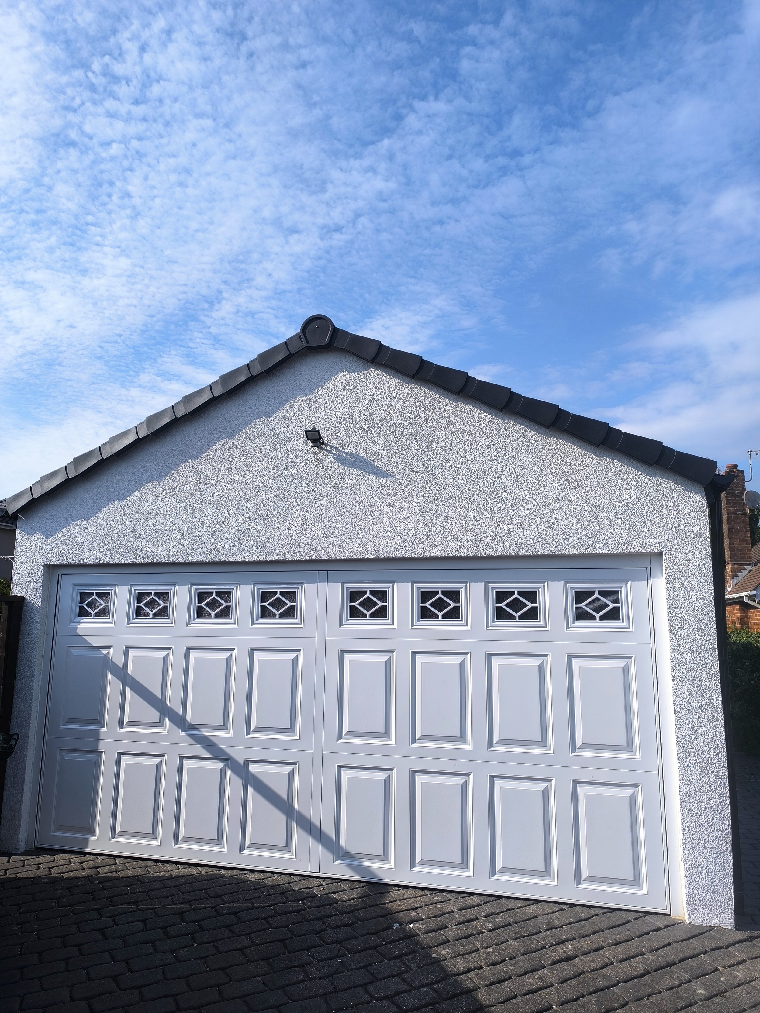 White garage door with small windows near the top, attached to a building with textured white walls and a dark tiled roof. The sky is blue with scattered clouds, and a shadow of a pole is cast on the garage door.