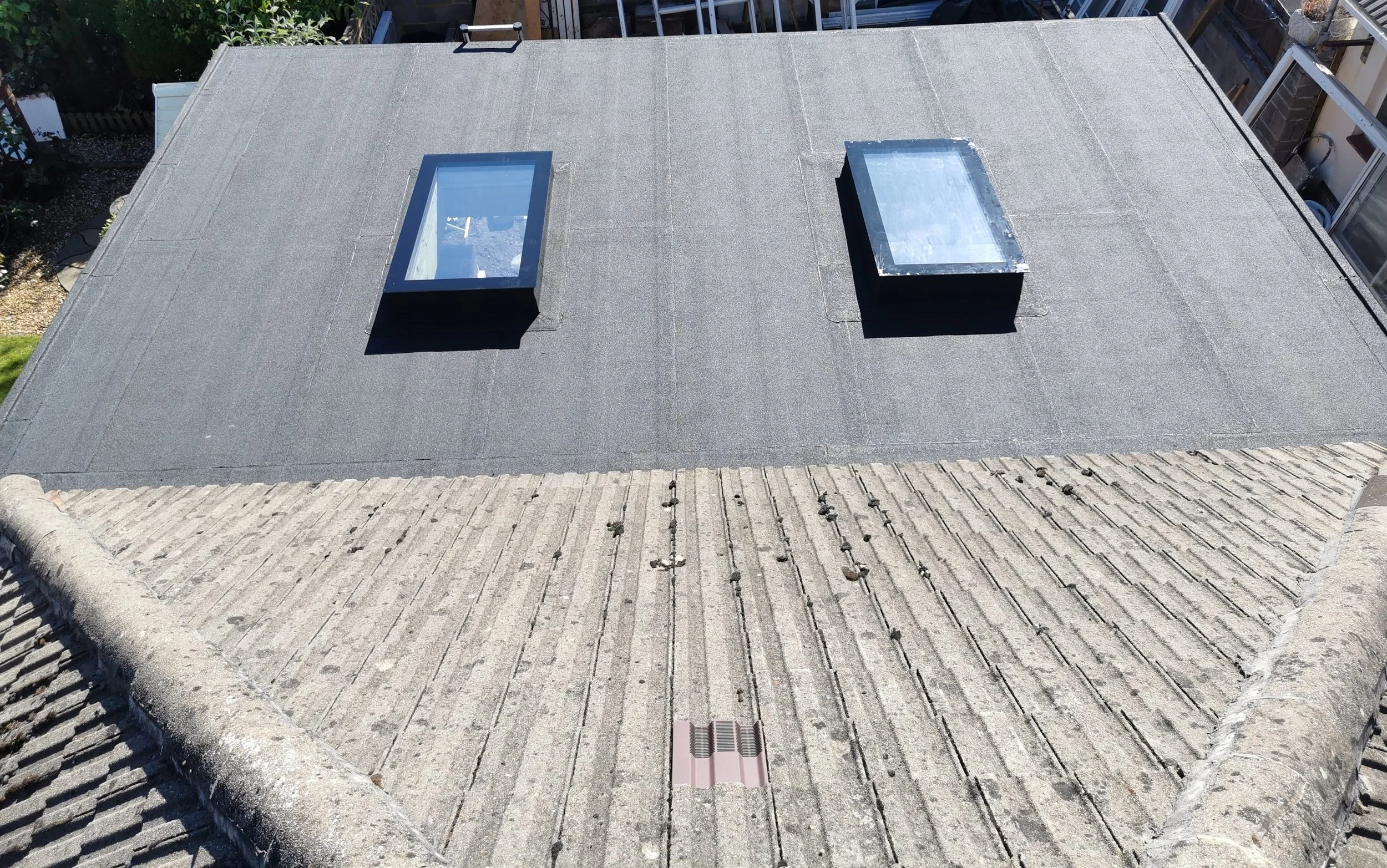 View of a roof with two skylights, one rectangular and one square, installed on a flat black roofing surface. The foreground shows a sloped tiled roof edge.