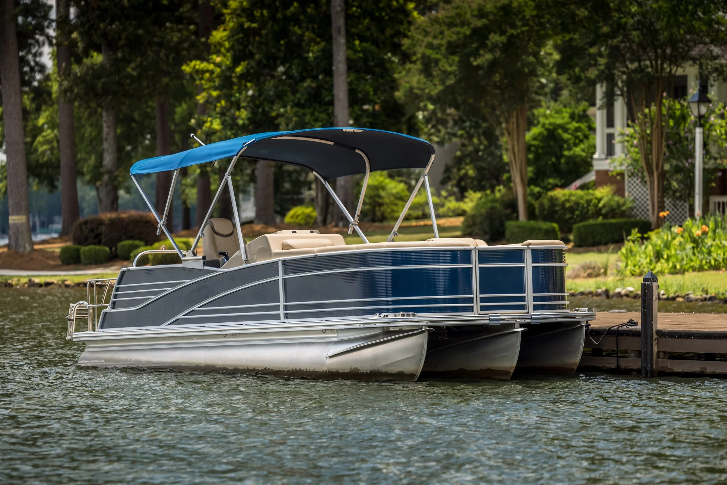 A pontoon boat docked at a wooden pier on a lake, with trees and a house in the background.