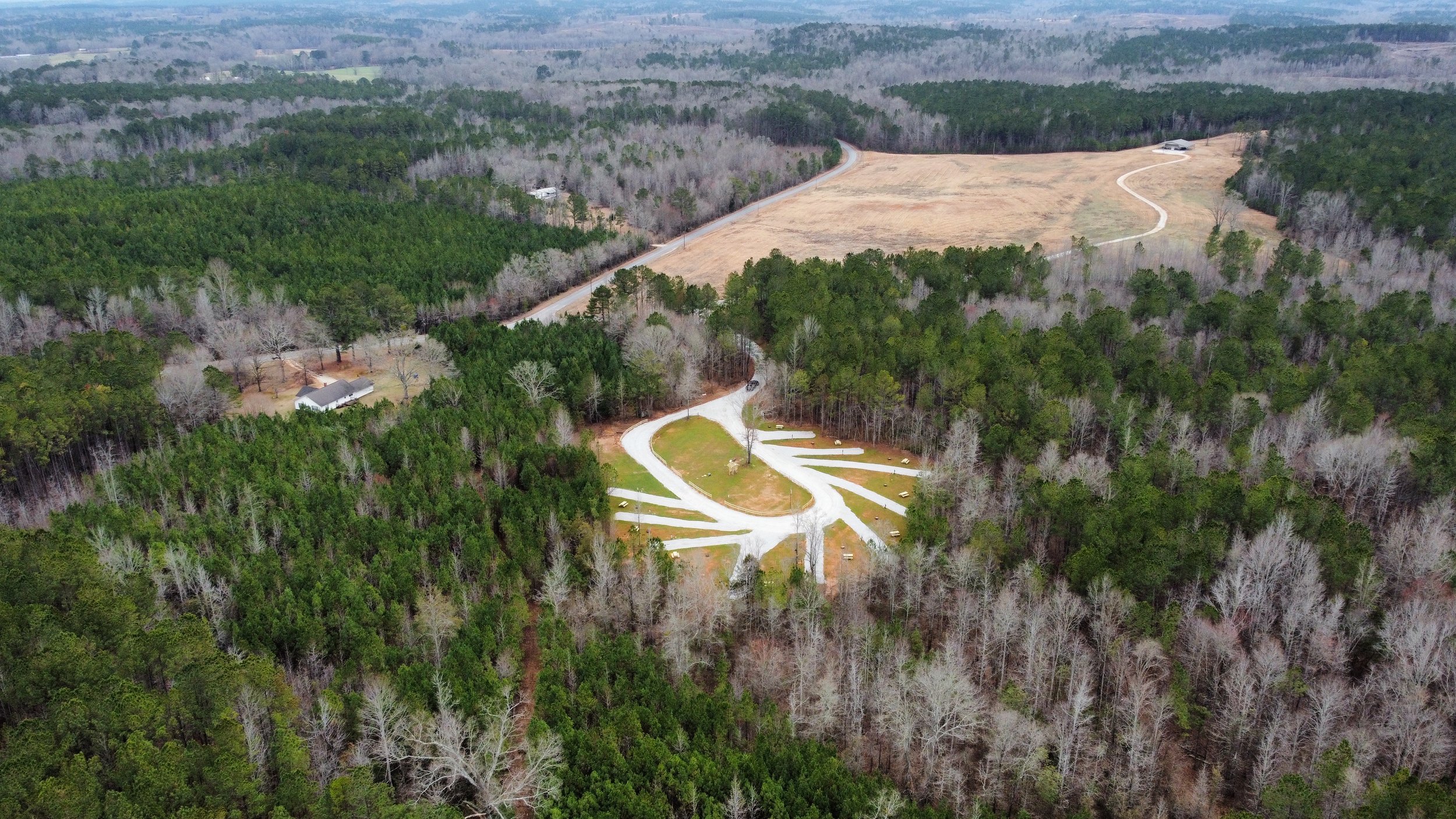 An aerial view of a rural landscape with a winding road, a clearing with a cemetery, dense trees, and some houses in the distance.