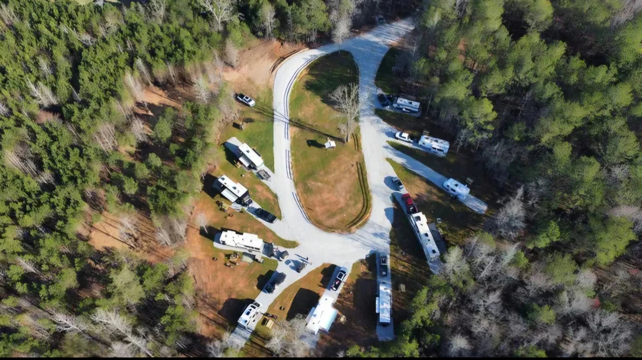 An aerial view of the campground surrounded by trees, with several RVs and vehicles parked around the campground and a winding gravel road passing through the area.