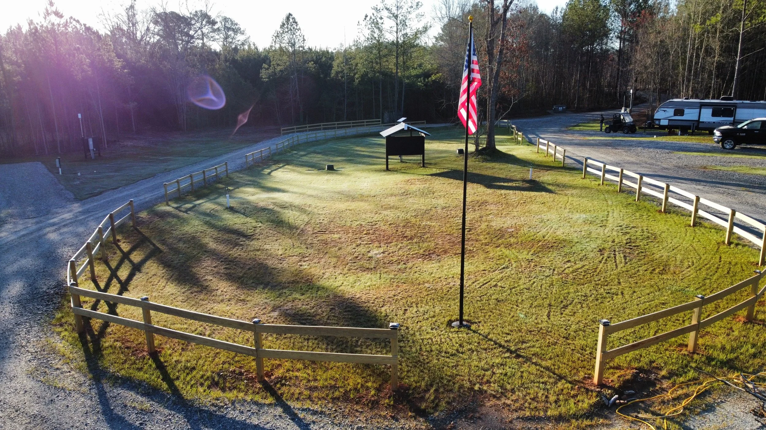 A grassy area surrounded by a wooden fence, with a flagpole and an American flag in the center. There is a small black structure, possibly a sign or bulletin board, near the flagpole. In the background, there are parked vehicles and trees, with some vehicles in what appears to be a parking lot.